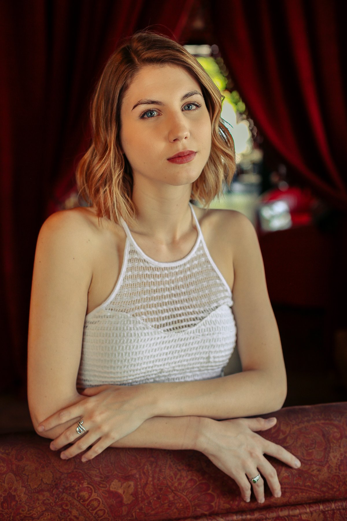 A young woman with shoulder-length wavy hair and blue eyes sitting at a table in a room with red curtains and greenery outside, wearing a white sleeveless top.