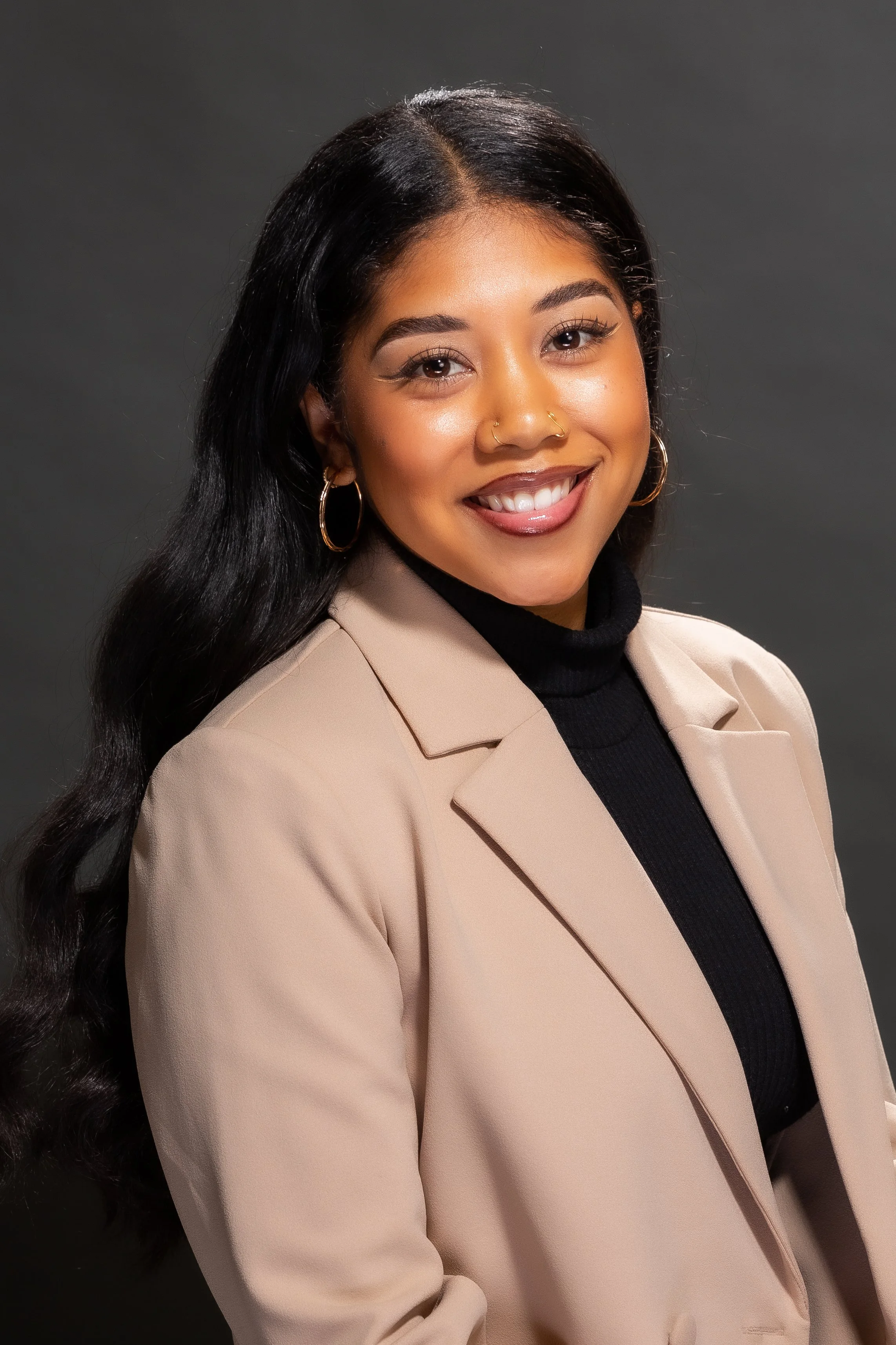 A young woman with long dark hair, wearing a beige blazer over a black turtleneck, smiling against a dark background.