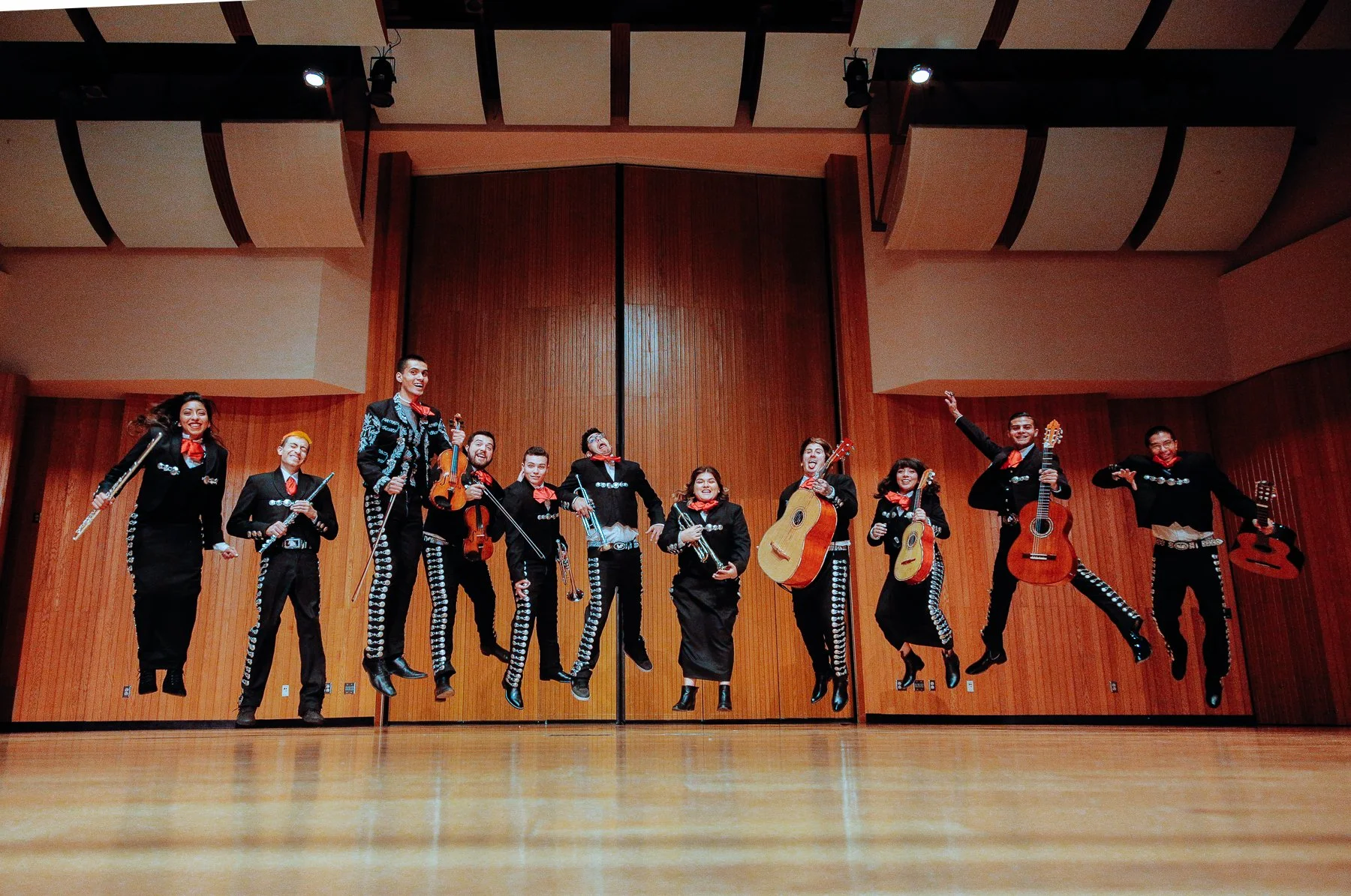 Ten musicians in Mariachi costumes jumping and holding musical instruments on a stage with wooden walls.