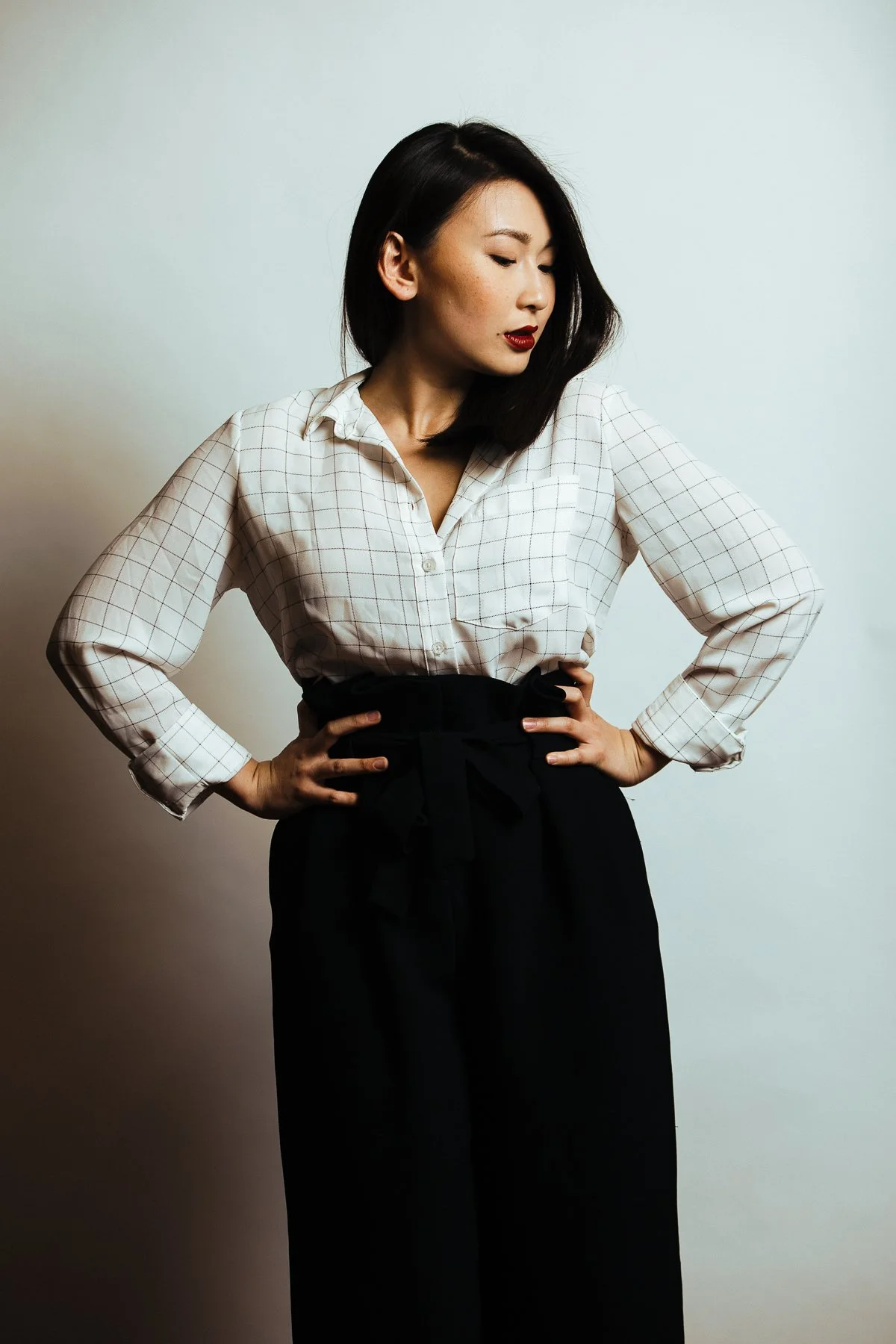 A woman with shoulder-length dark hair, wearing a white checkered shirt tucked into black high-waisted trousers, standing with her hands on her hips against a plain light-colored background, looking down and to the side.