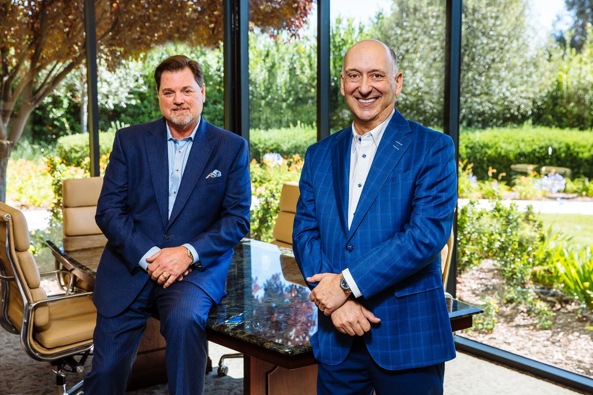 Two men in business suits standing in a conference room with large windows overlooking a garden. One man is sitting on the edge of a desk, and the other is standing, both smiling.