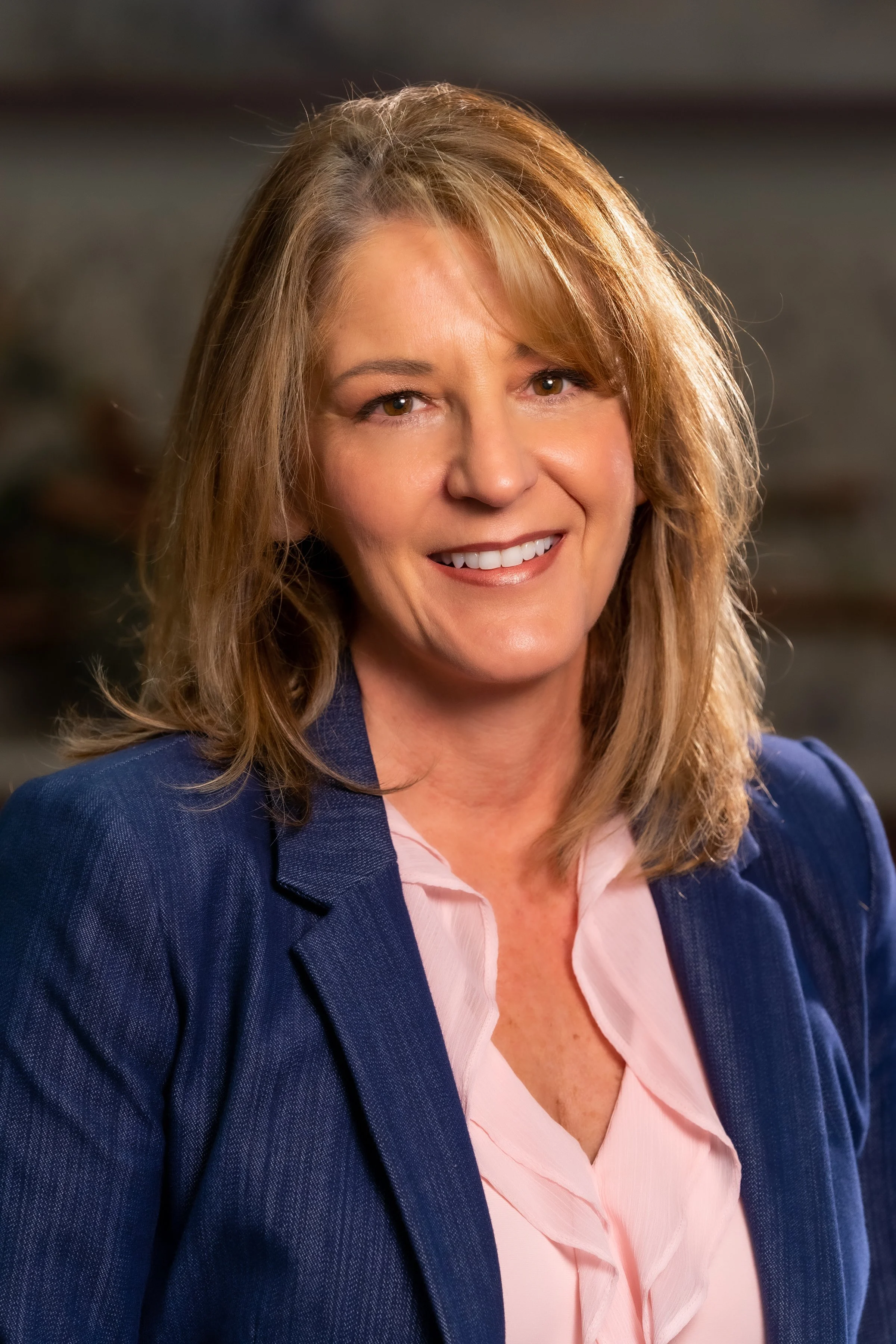 A smiling woman with shoulder-length blonde hair, wearing a dark blue blazer and a light pink blouse, in an indoor setting with a blurred background.