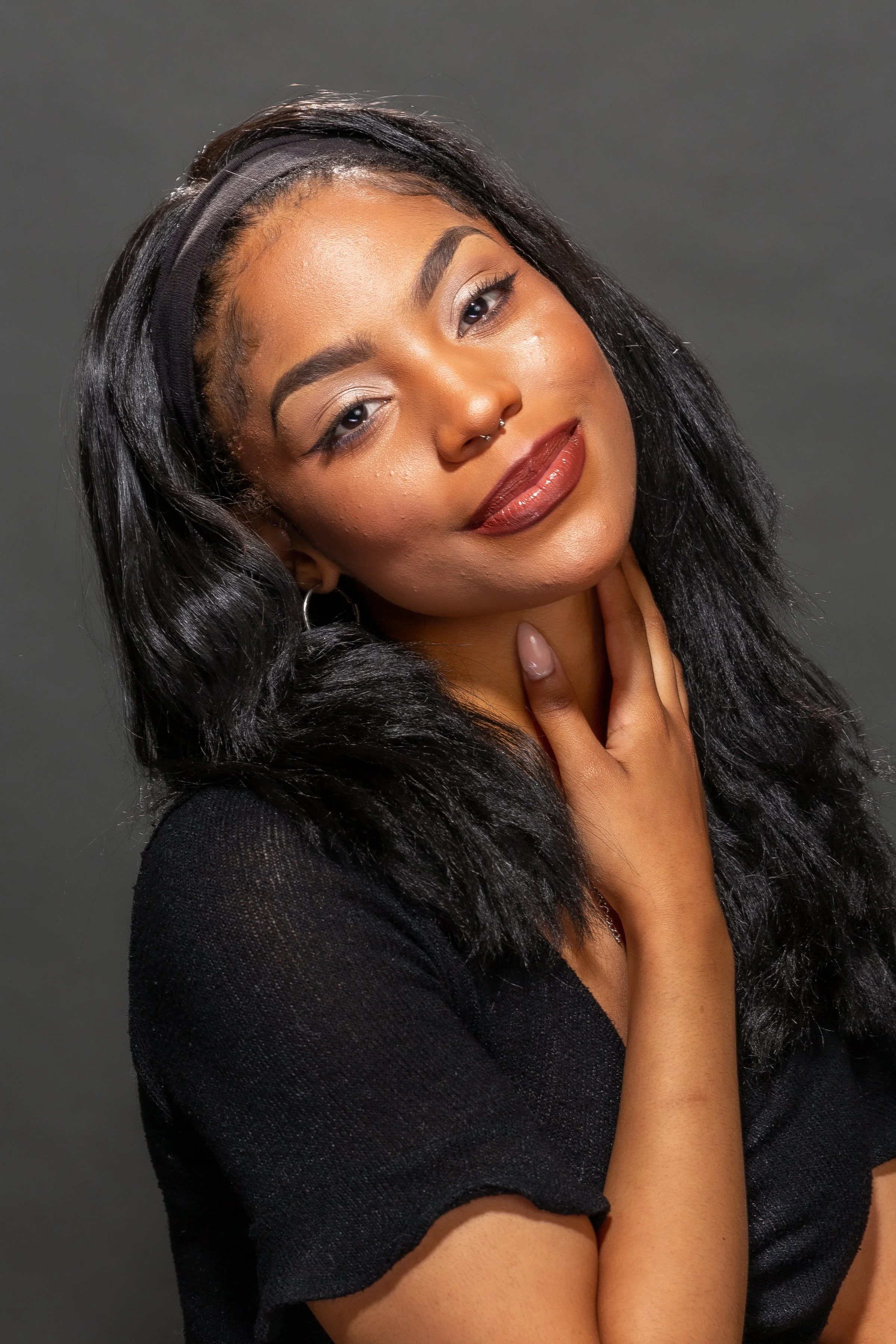A young woman with long black hair, wearing a black headband and black top, poses with her hand on her neck, smiling softly against a dark background.