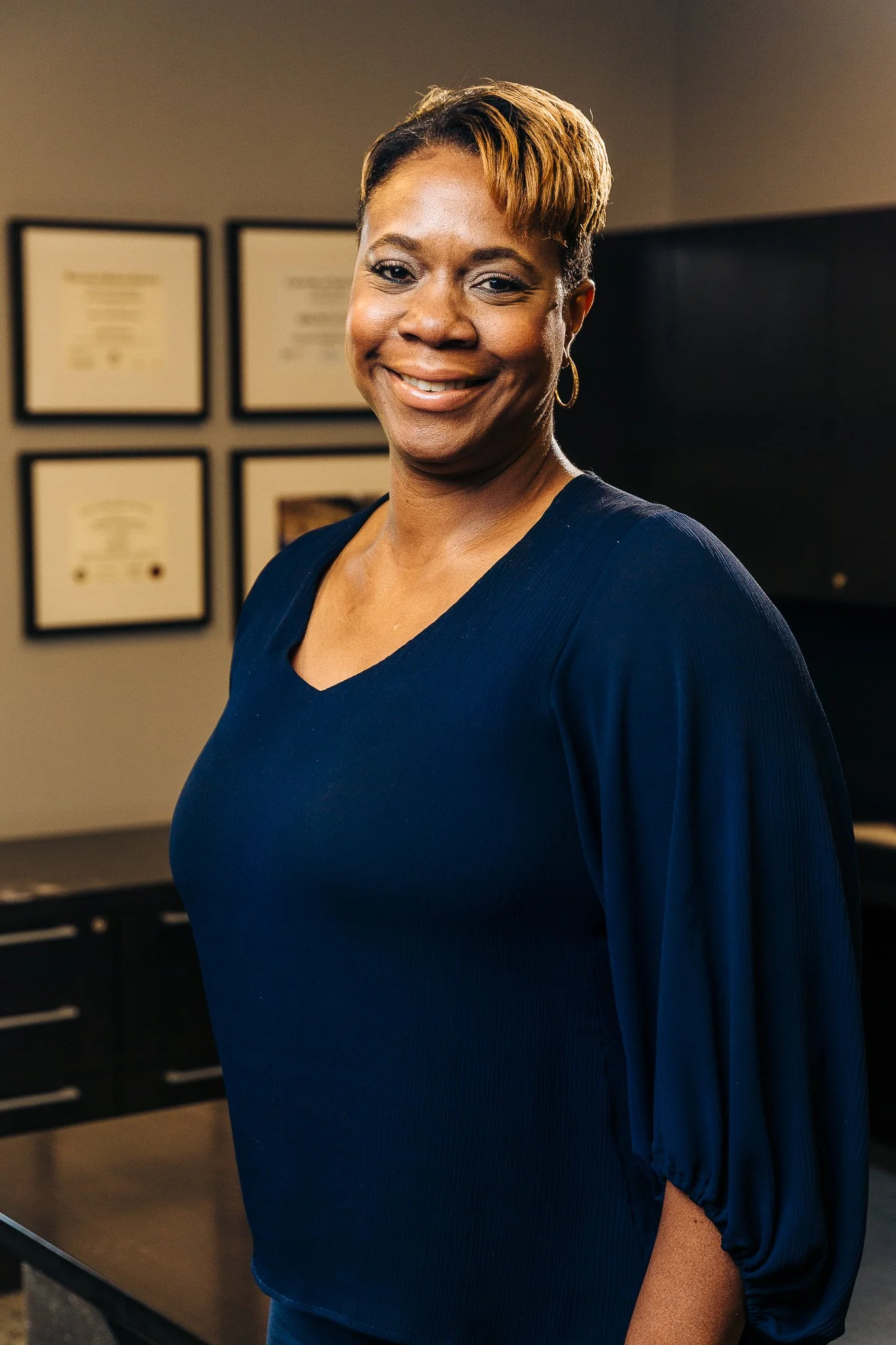 A smiling woman with short hair, wearing a navy blue top with puffed sleeves, standing in an office or professional setting with framed certificates or diplomas on the wall behind her.