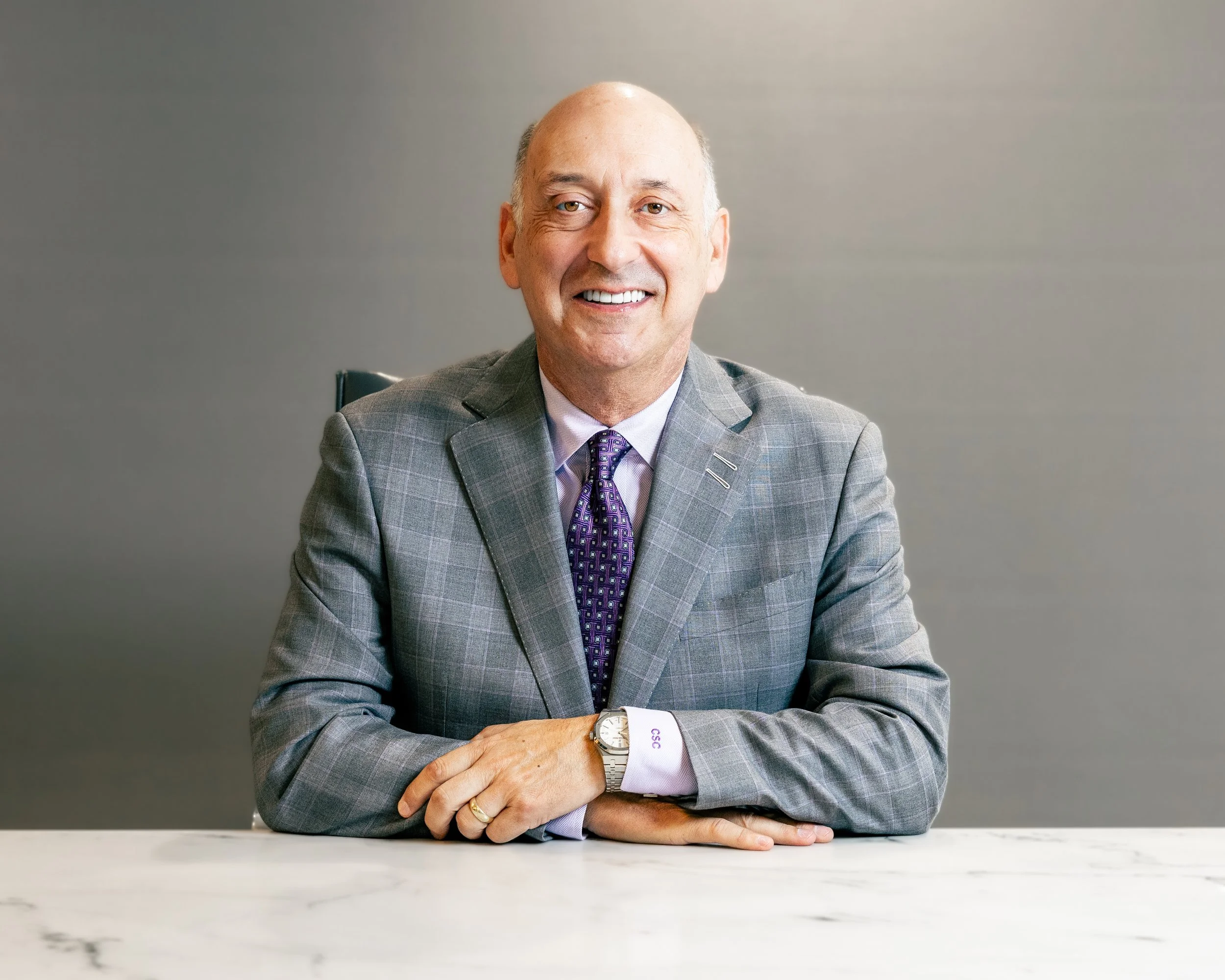 A smiling middle-aged man in a gray plaid suit, purple shirt, and tie, sitting at a white marble table with his hands folded, in front of a plain gray background.