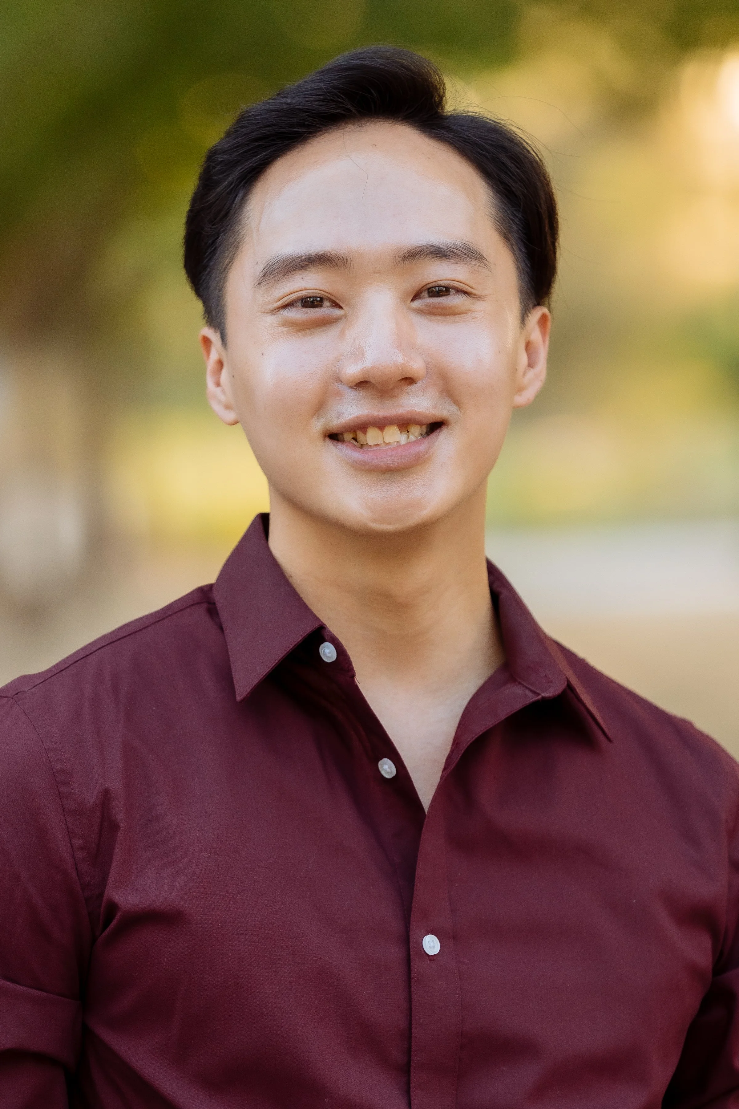 A young man smiling outdoors, wearing a maroon button-up shirt, with a blurred green and yellow background.