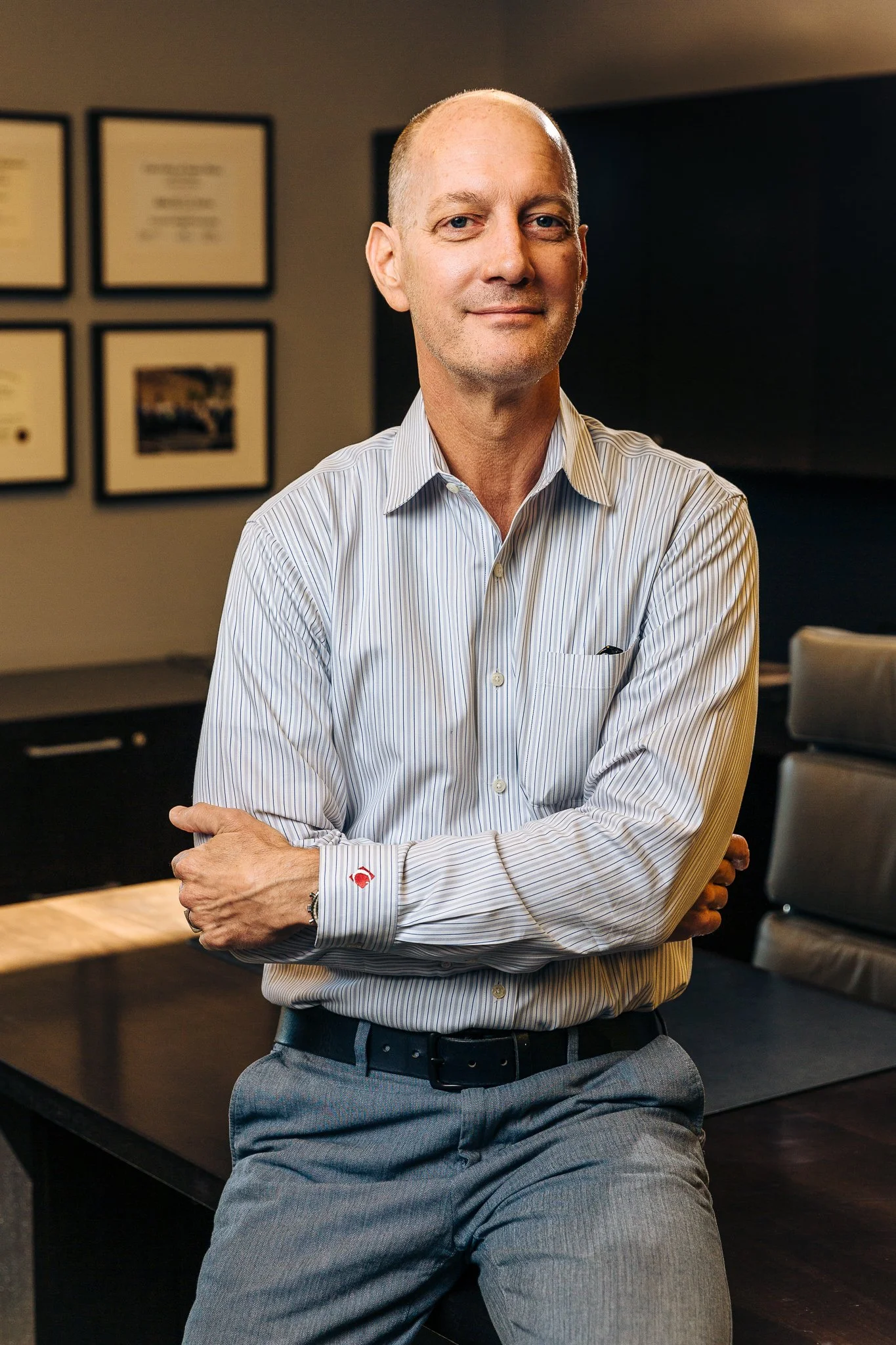 A middle-aged man with a shaved head and light skin, wearing a light blue striped dress shirt and gray pants, standing in a conference room with framed documents on the wall, arms crossed, looking at the camera.