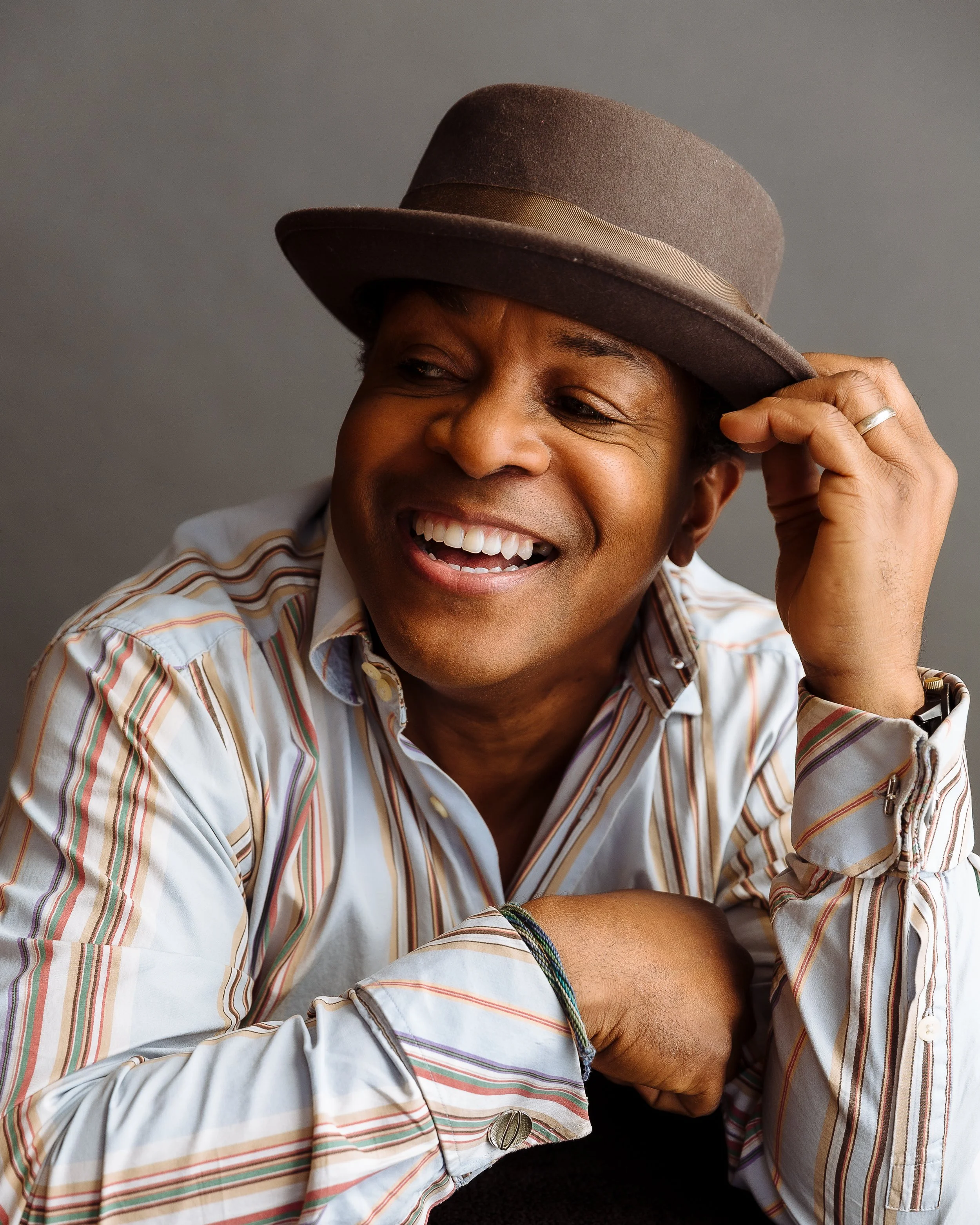 A man wearing a striped shirt and a brown hat, smiling and touching his hat, against a gray background.