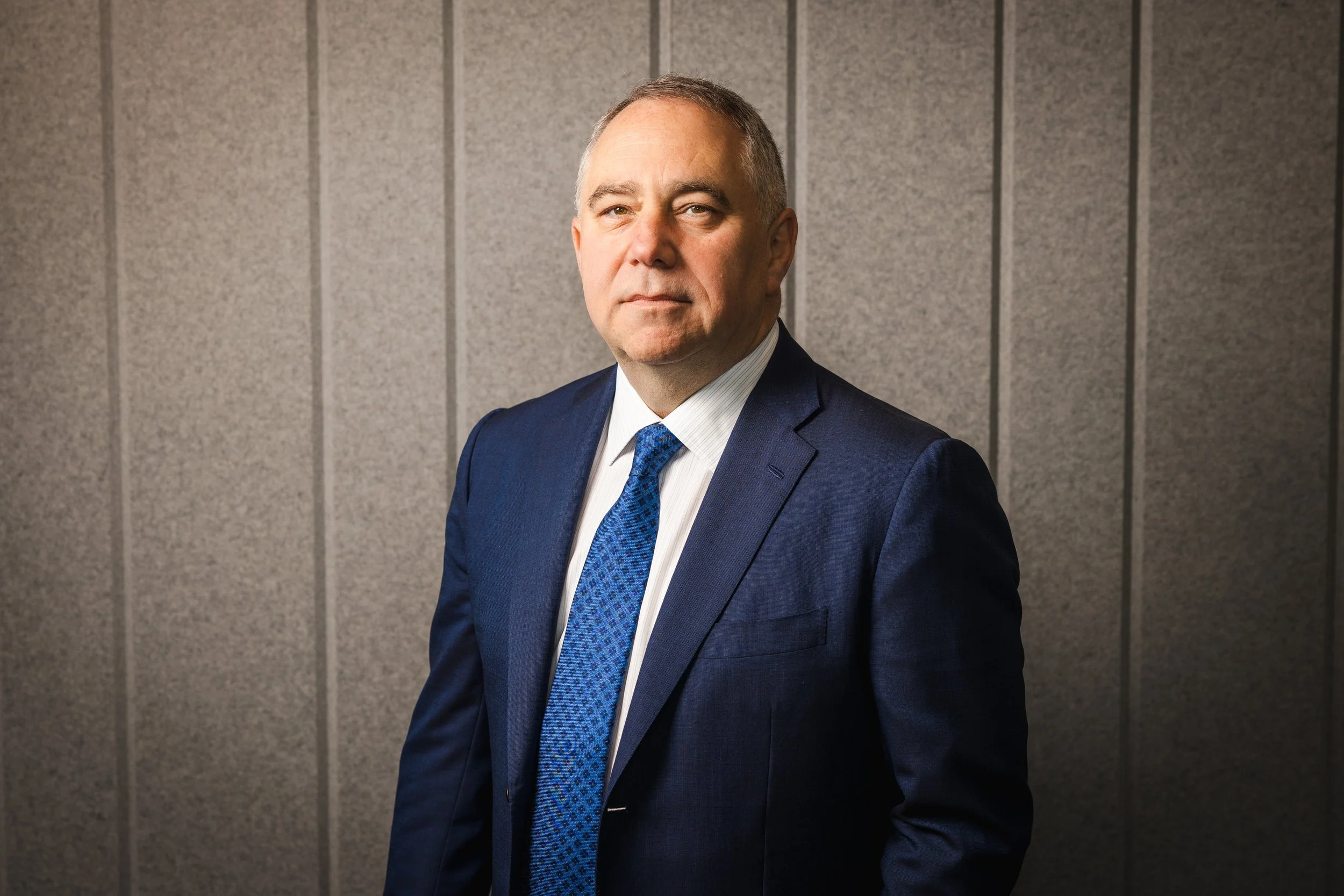 A middle-aged man in a dark blue suit, white shirt, and blue patterned tie standing against a textured gray wall.