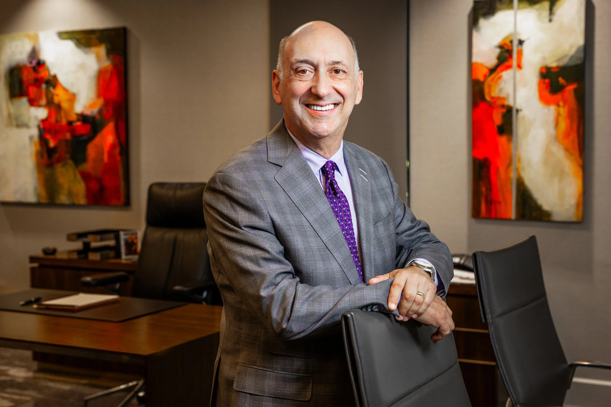 A smiling middle-aged man in a gray suit with a purple tie, standing in a modern office conference room with abstract art on the walls, leaning on a black leather chair.