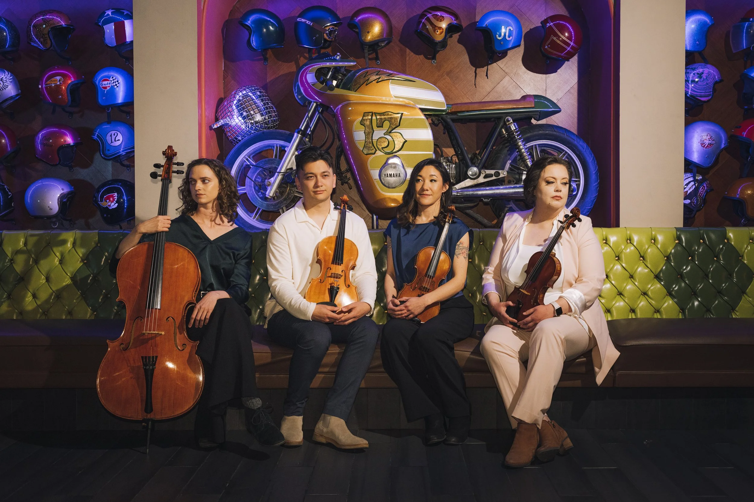 Four women and one man seated on a green tufted bench, each holding a violin in a room decorated with vintage motorcycle helmets and a yellow motorcycle in the background.