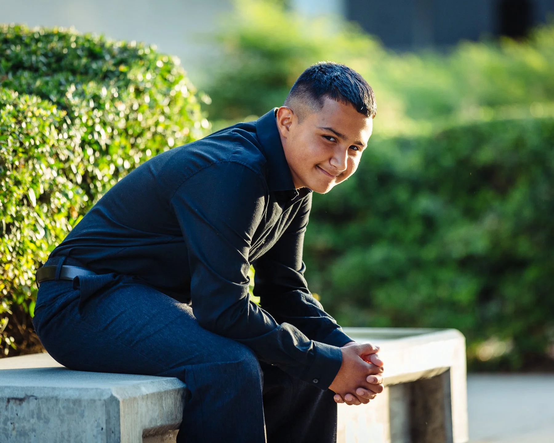 A young man with short dark hair, wearing a black shirt and dark jeans, sitting on a concrete bench outdoors, smiling and looking at the camera, with greenery in the background.