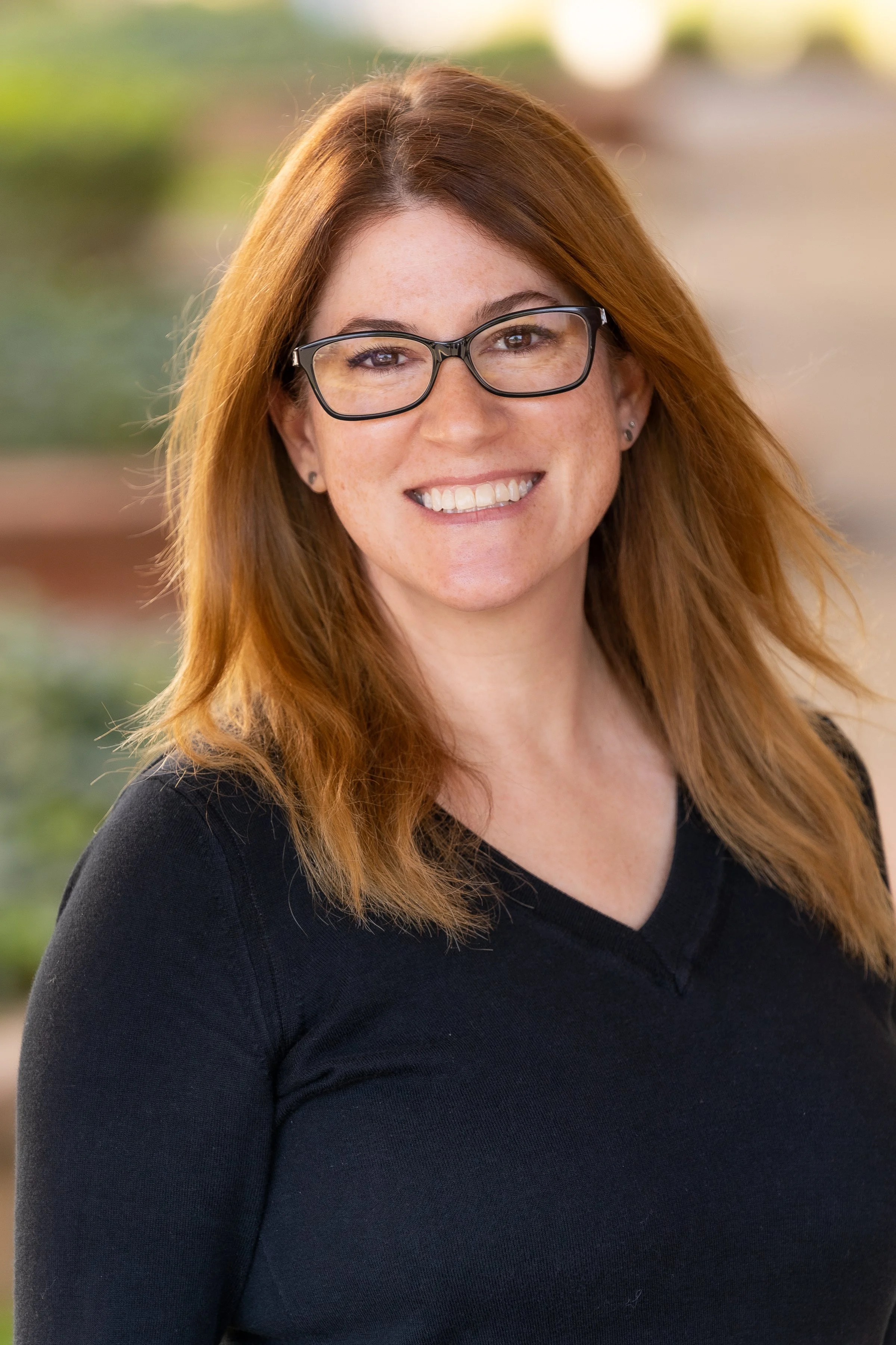 A woman with long red hair, wearing glasses and a black shirt, smiling outdoors with a blurred background.