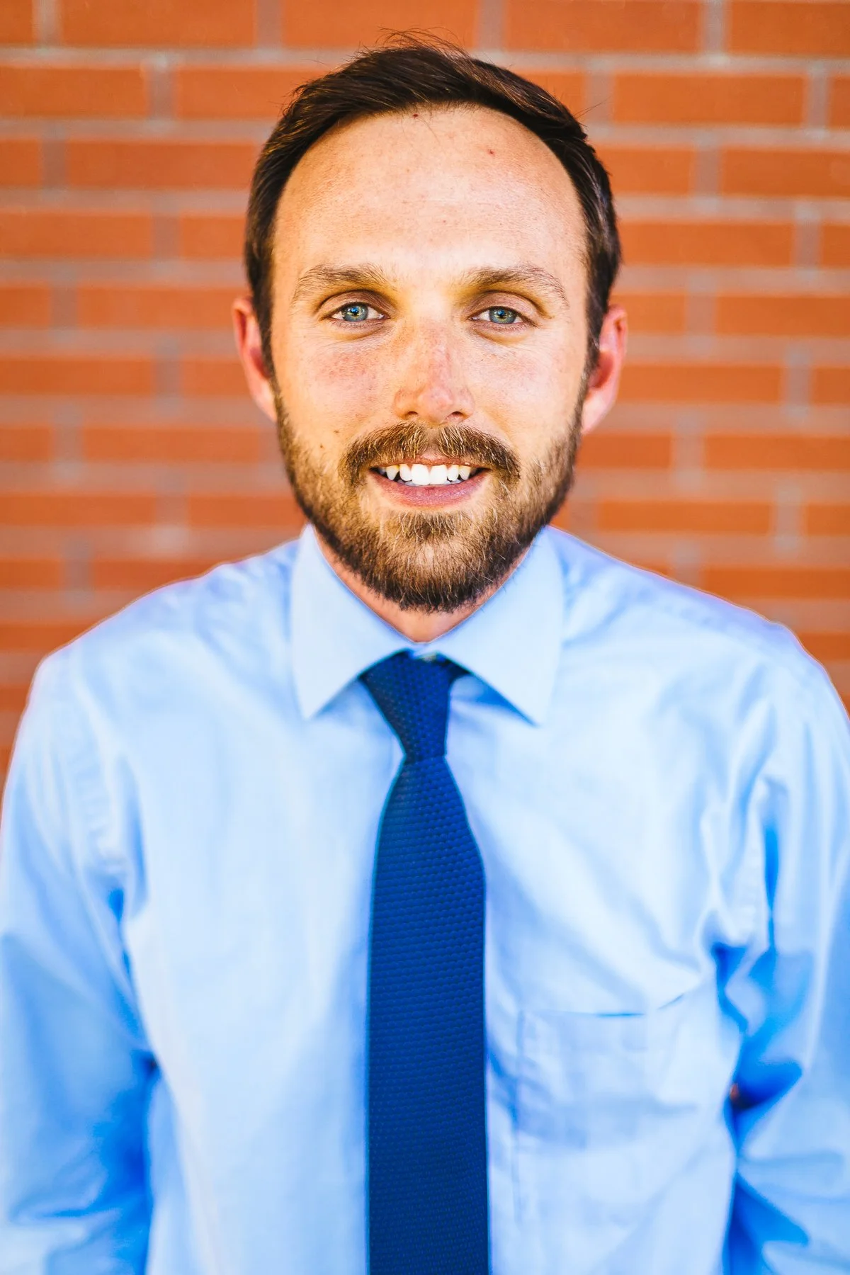 Smiling man in a blue dress shirt and dark blue tie, standing in front of a brick wall.