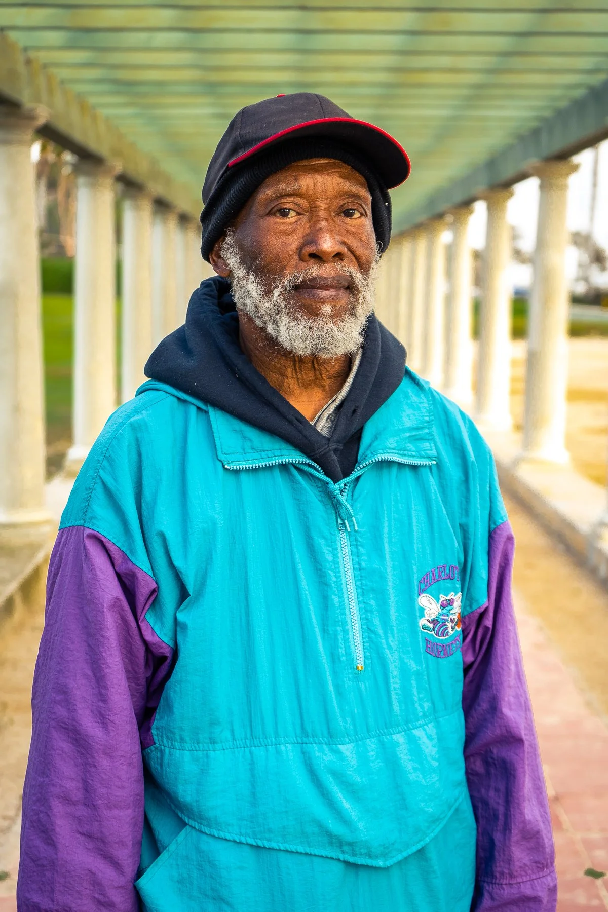 An elderly man with gray beard and dark skin wearing a black and red baseball cap, a black hoodie, and a colorful blue and purple windbreaker jacket standing outdoors under a colonnade.