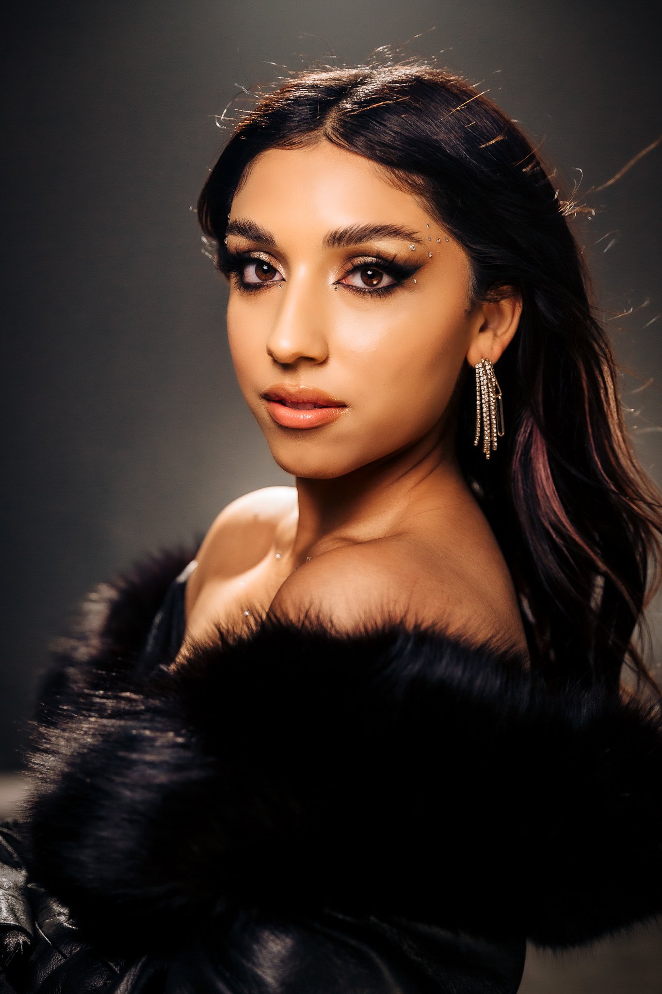 A woman with long dark hair, wearing jewelry and a fur coat, looking at the camera with a confident expression against a dark background.