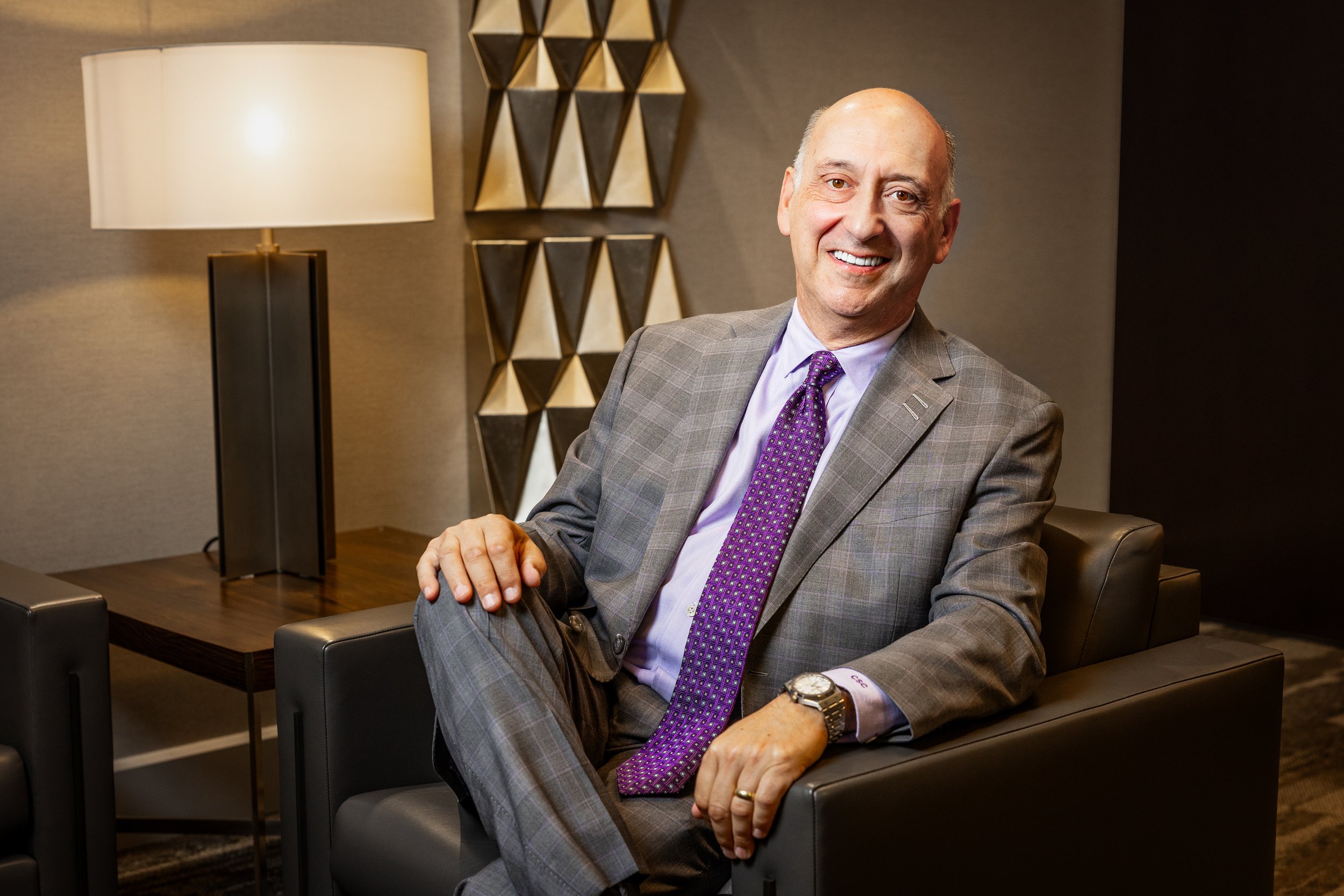 A smiling man in a gray checkered suit and purple tie sitting on a black chair in a modern office or lounge with a decorative wall art, a wooden table, and a table lamp in the background.