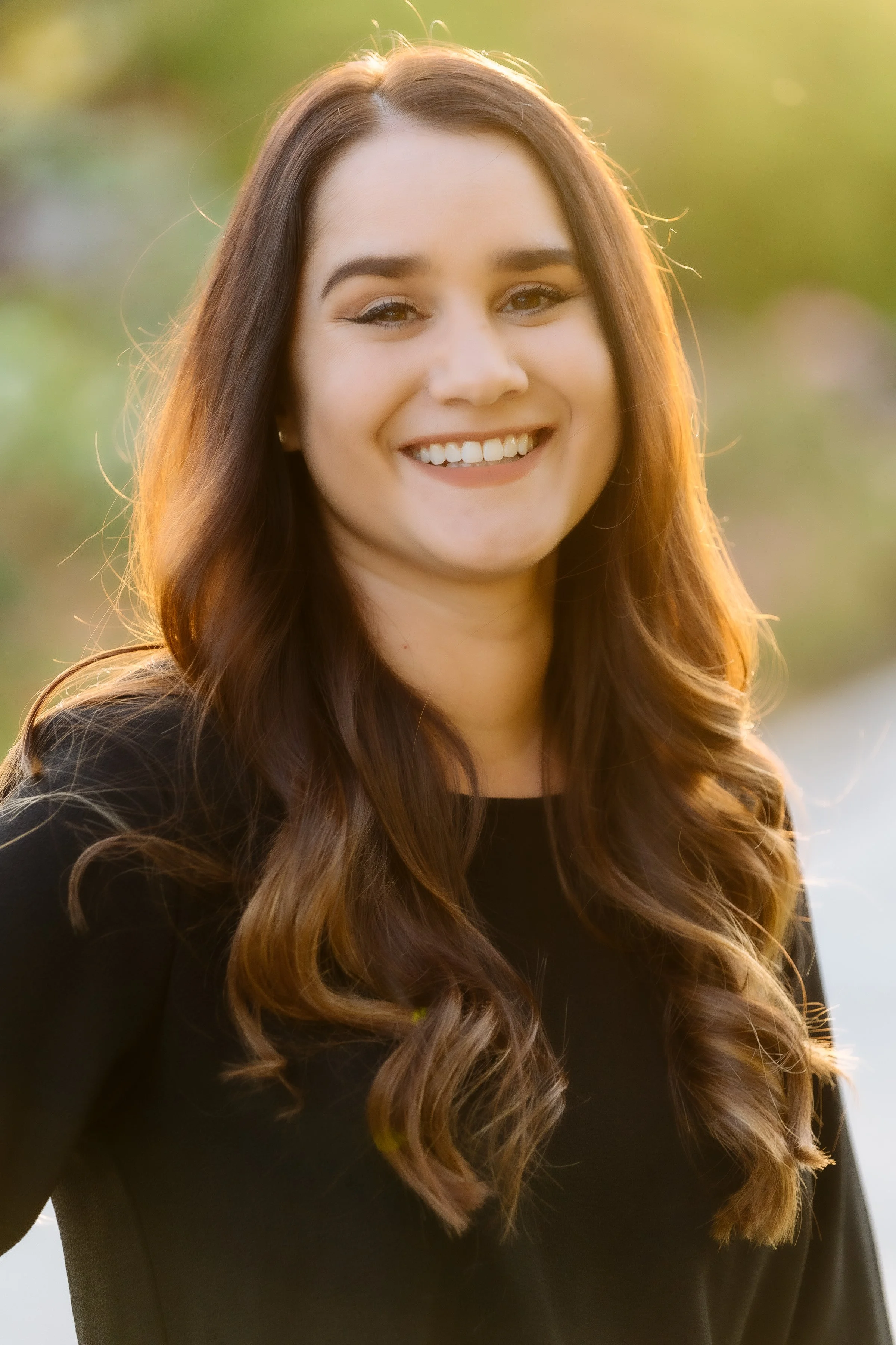 Portrait of a smiling young woman with long wavy brown hair, wearing a black top, outdoors during sunset with blurred natural background.