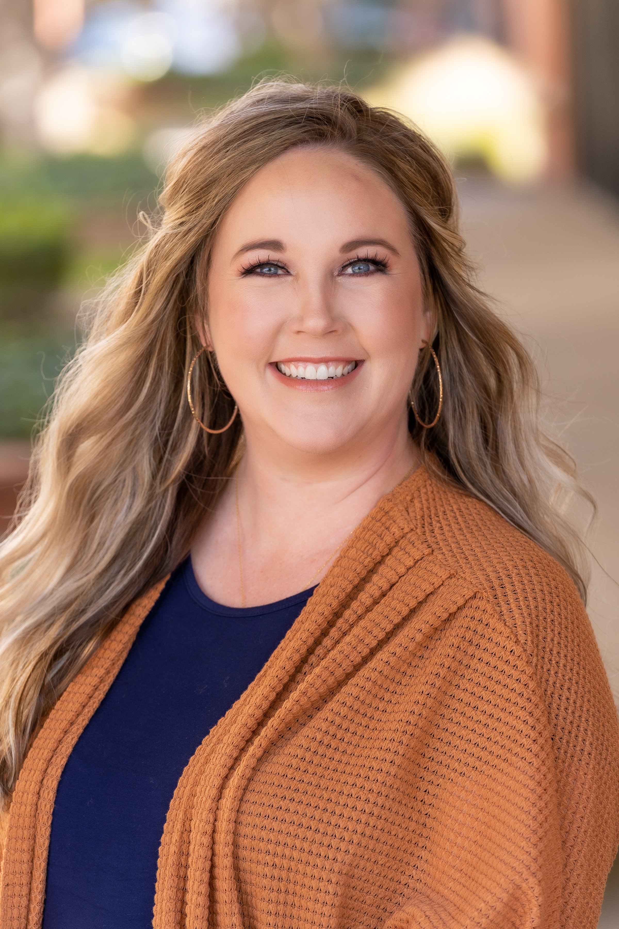 A woman with long wavy blonde hair smiling outdoors, wearing hoop earrings, a navy blue top, and an orange cardigan.