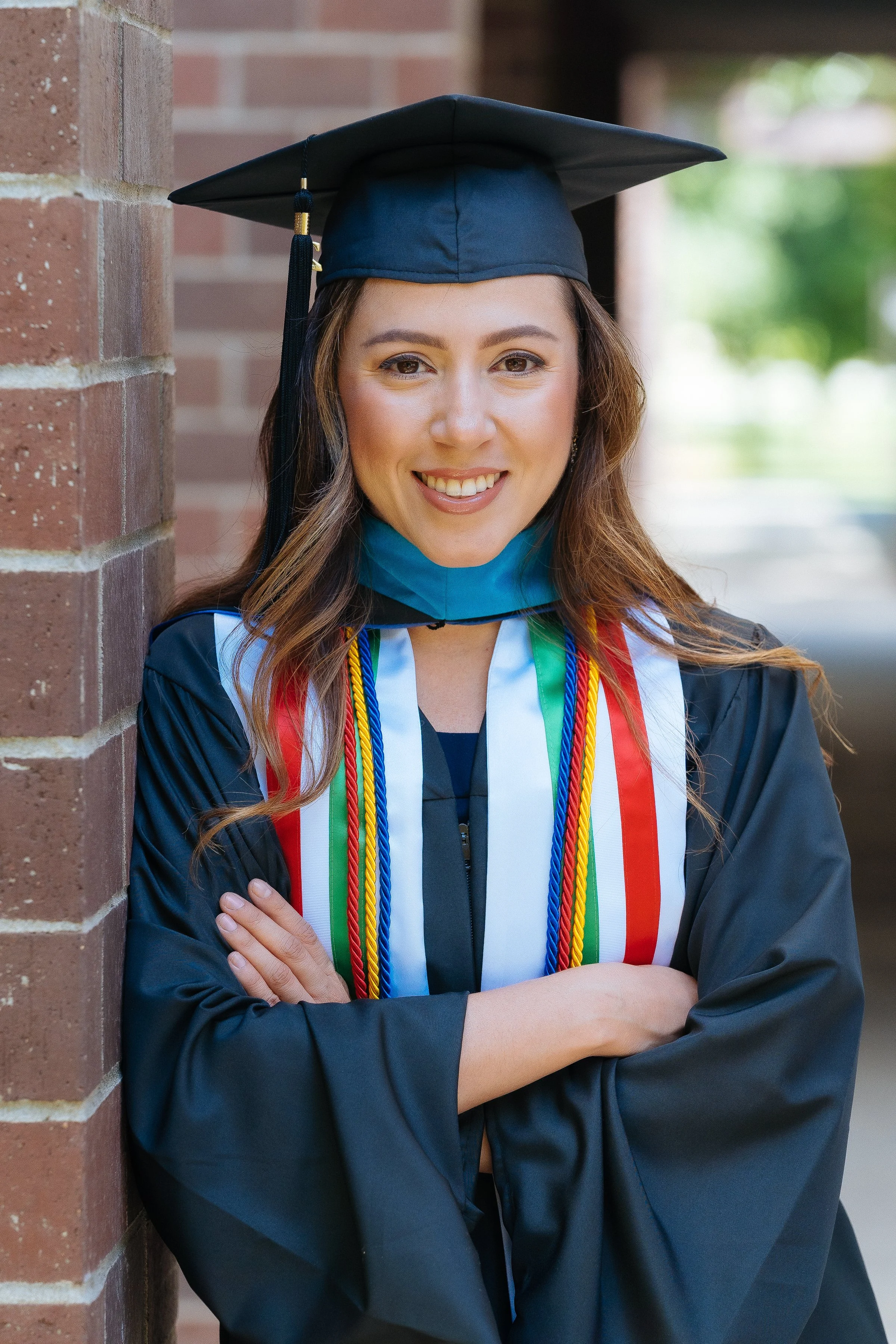 A female graduate in a cap and gown with medals and cords poses for a photo outside, leaning against a brick wall, with a bright smile.