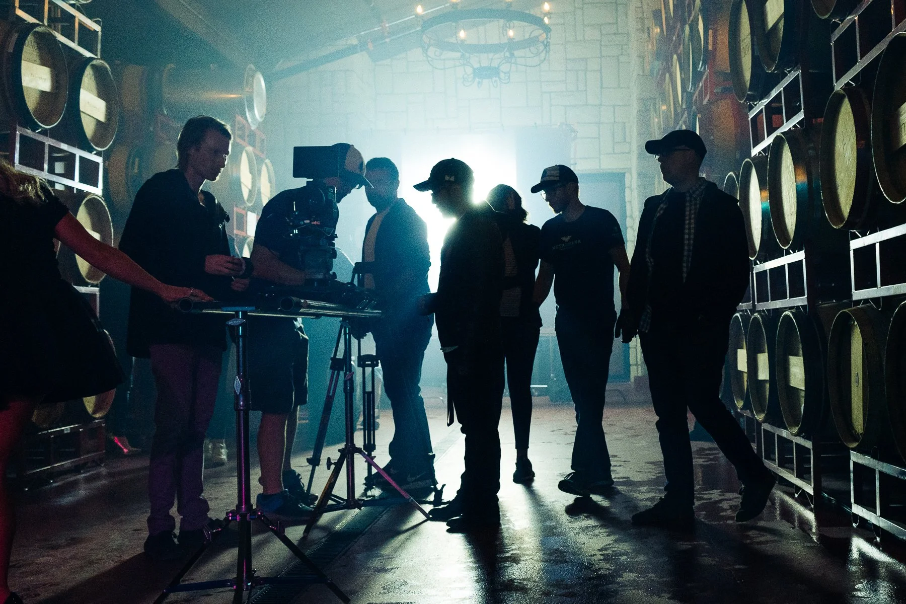 People working on a film set with a camera and crew members silhouetted against bright backlight in an industrial setting with large barrels on walls.