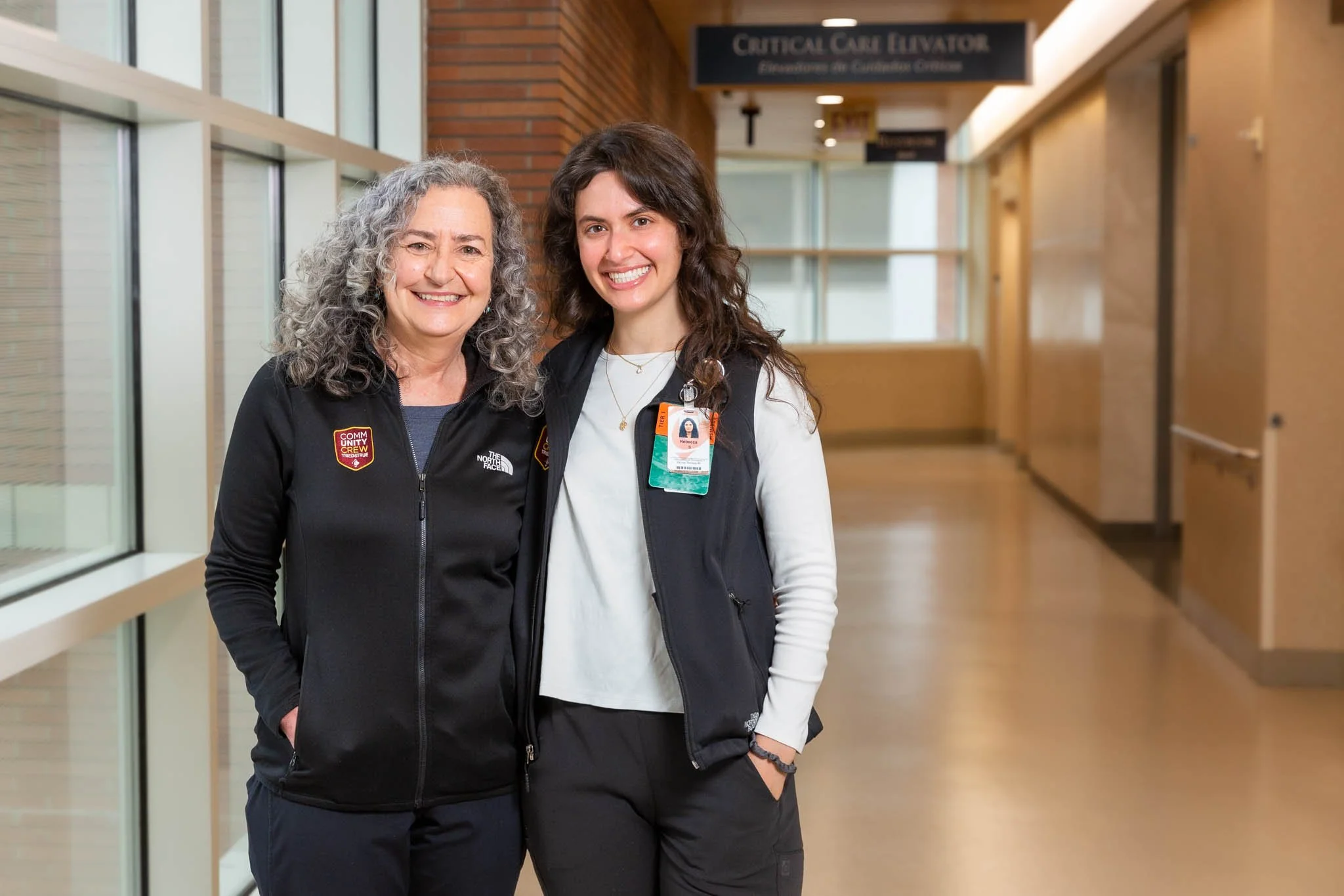 Two women smiling in hospital hallway, one with curly gray hair wearing a black jacket with patches, the other with wavy brown hair wearing a black vest, both with hospital badges.