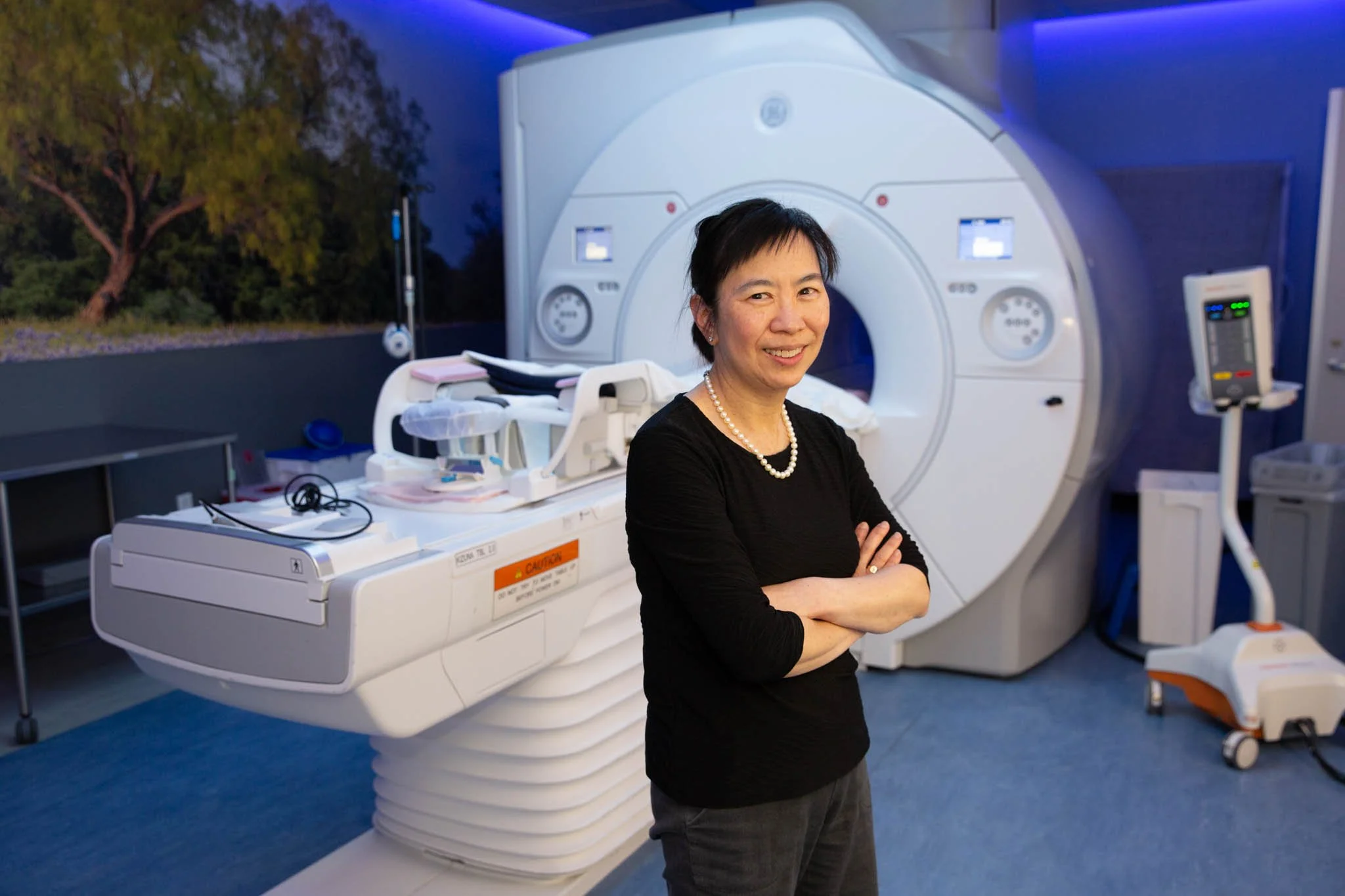A woman standing in front of an MRI machine in a medical facility, smiling with arms crossed.