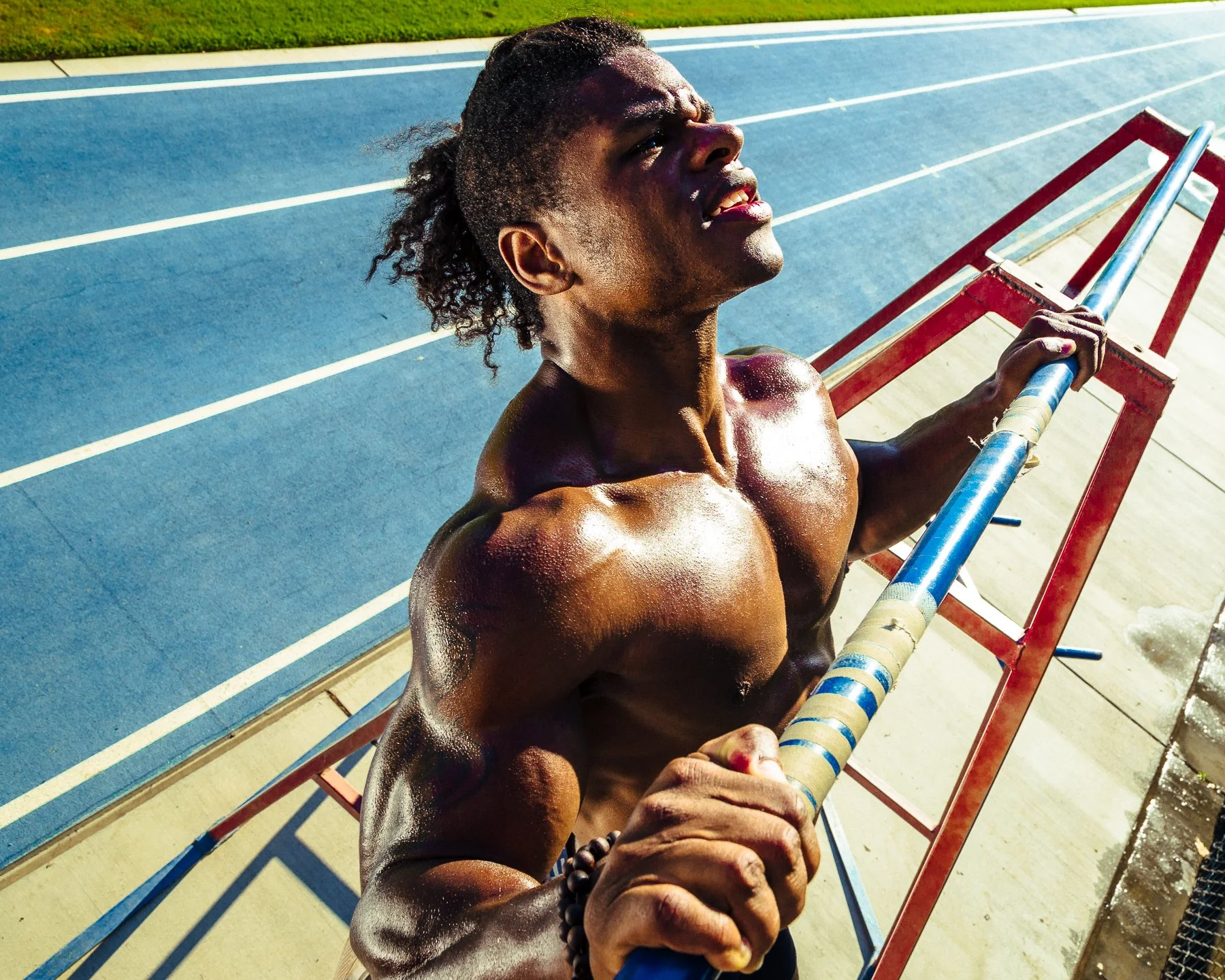 A male athlete is pushing himself during an outdoor workout, gripping a bar while using a ladder for support on a sunny day at a running track.