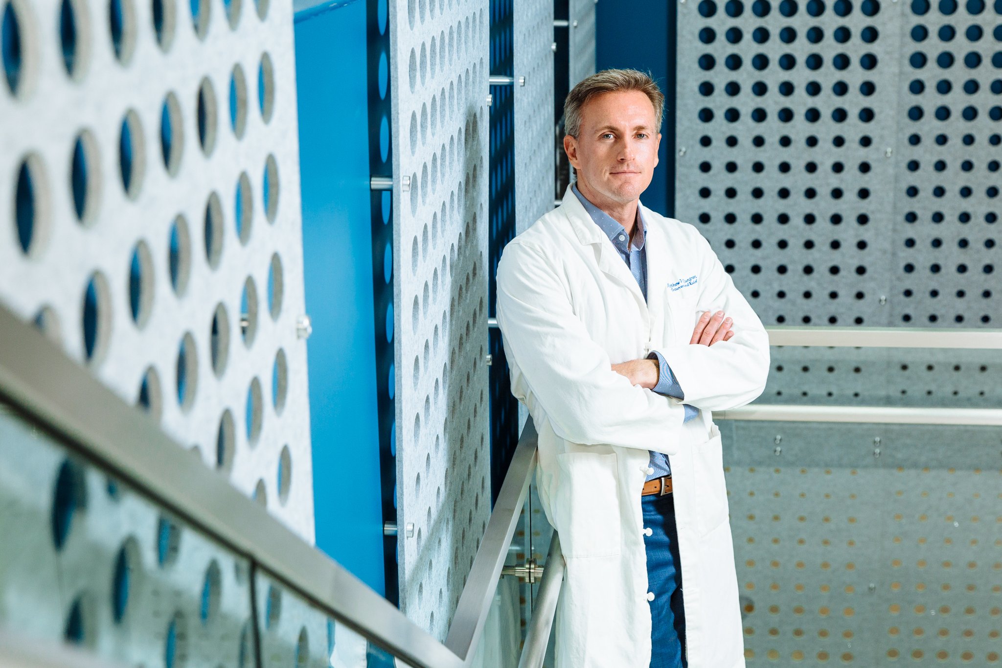 A male scientist or doctor standing with arms crossed in a laboratory or medical facility, with blue and gray perforated metal panels in the background.