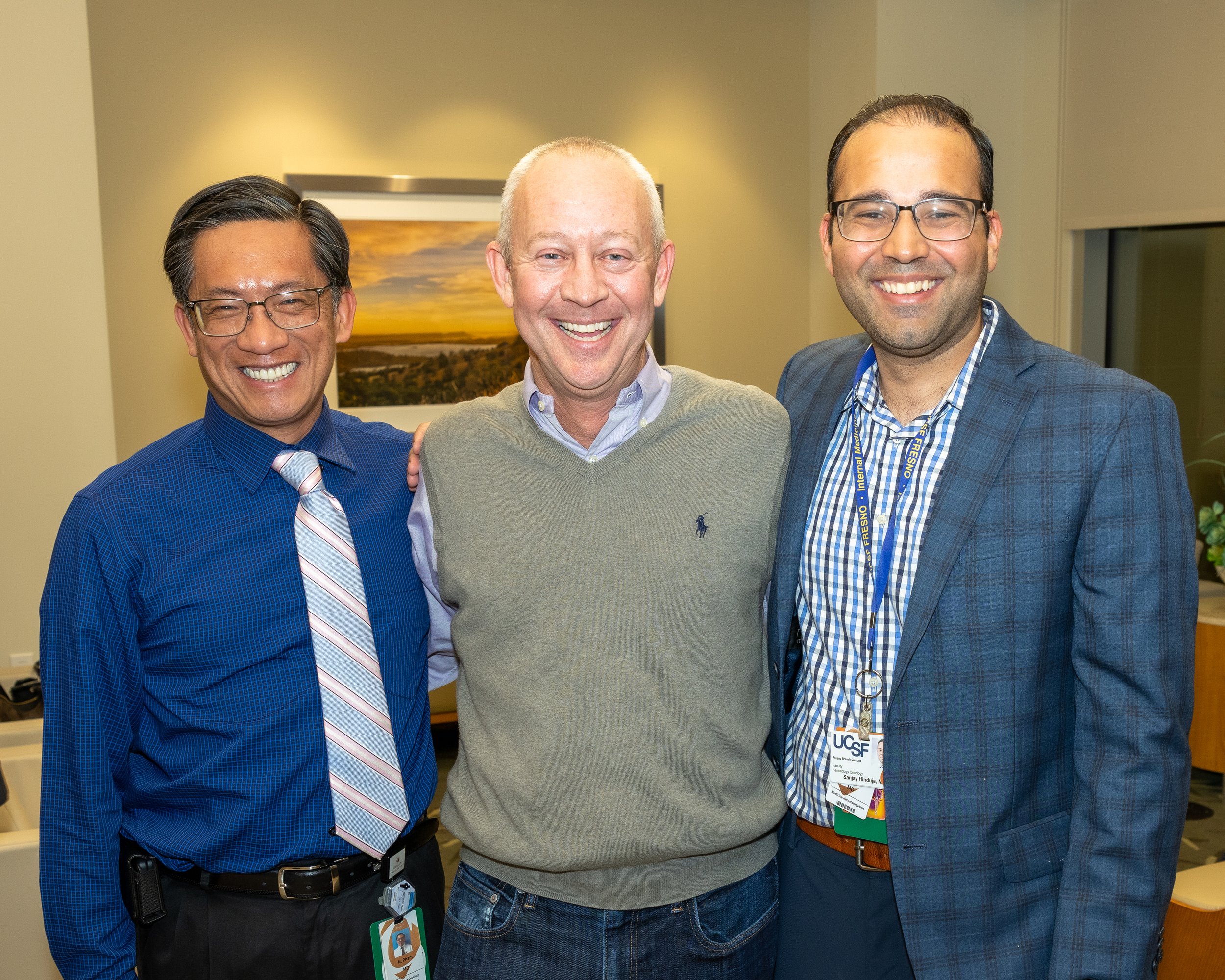 Three men smiling and standing close together indoors, with a framed landscape picture in the background. They are dressed in business casual attire.