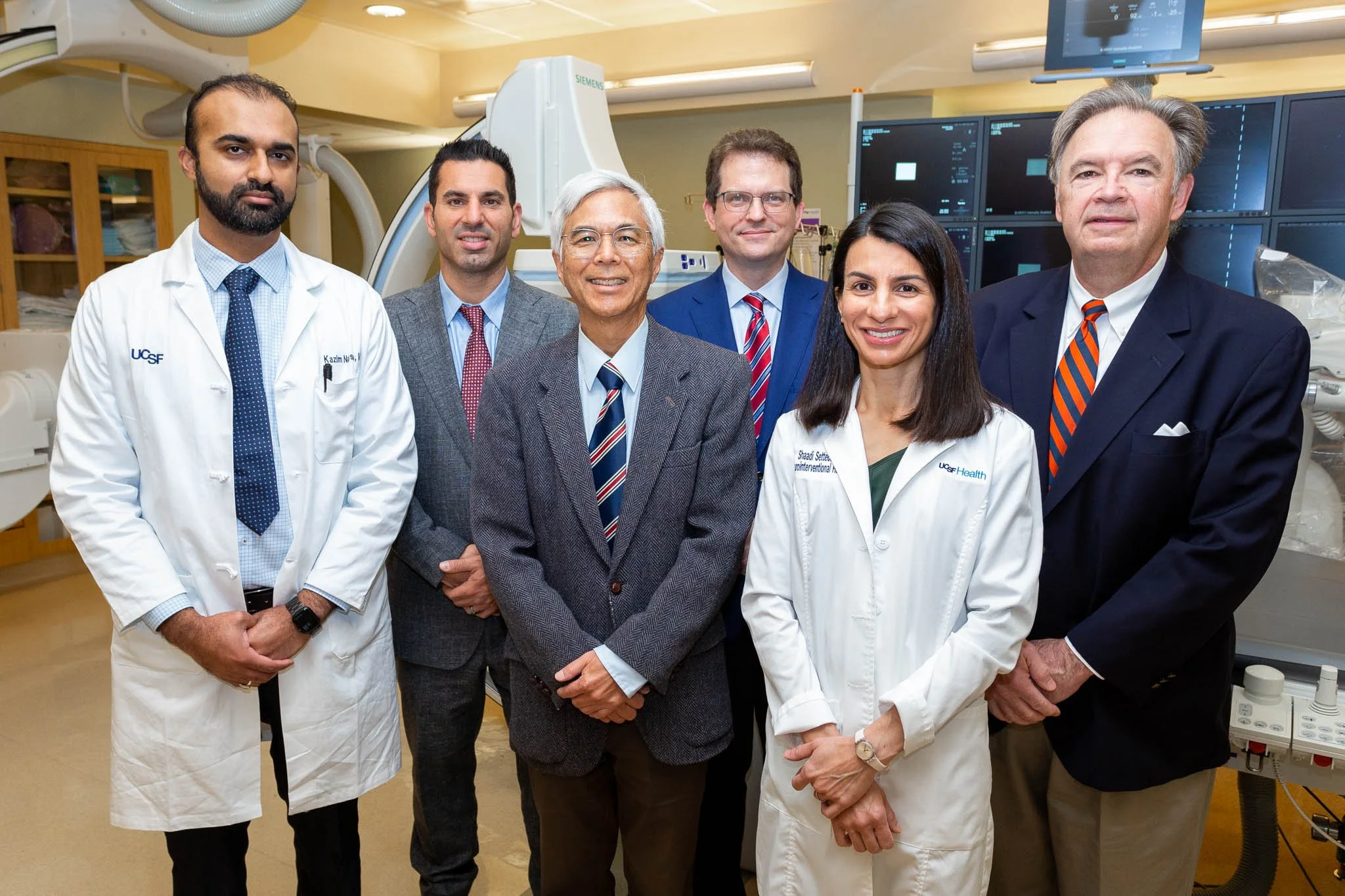 Group of seven diverse medical professionals and researchers in a hospital or laboratory setting, some in white coats and some in suits, standing together in front of medical equipment and monitors.