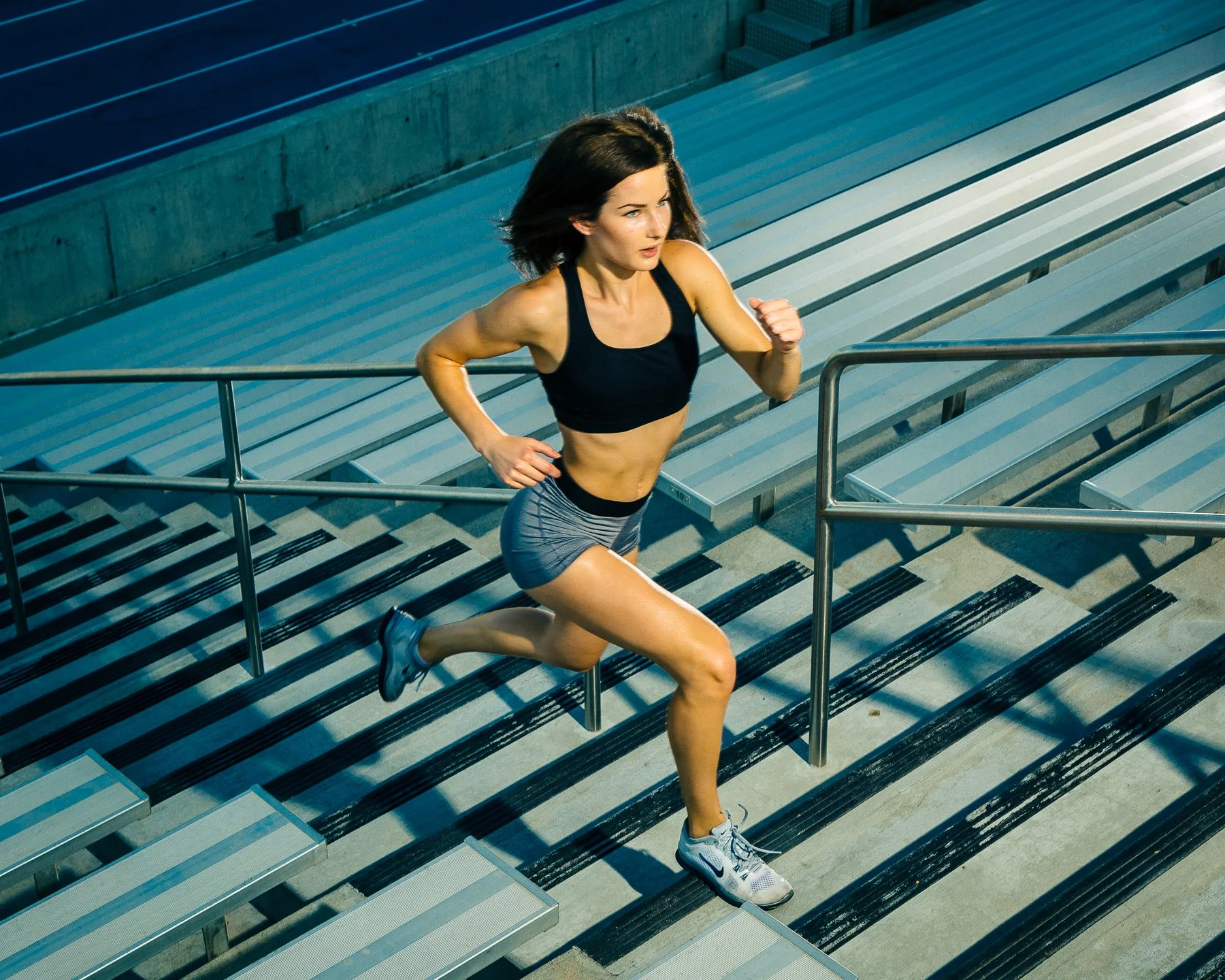 A woman running up outdoor stadium bleacher stairs with a handrail on her right side, wearing a black sports bra, gray shorts, and running shoes.