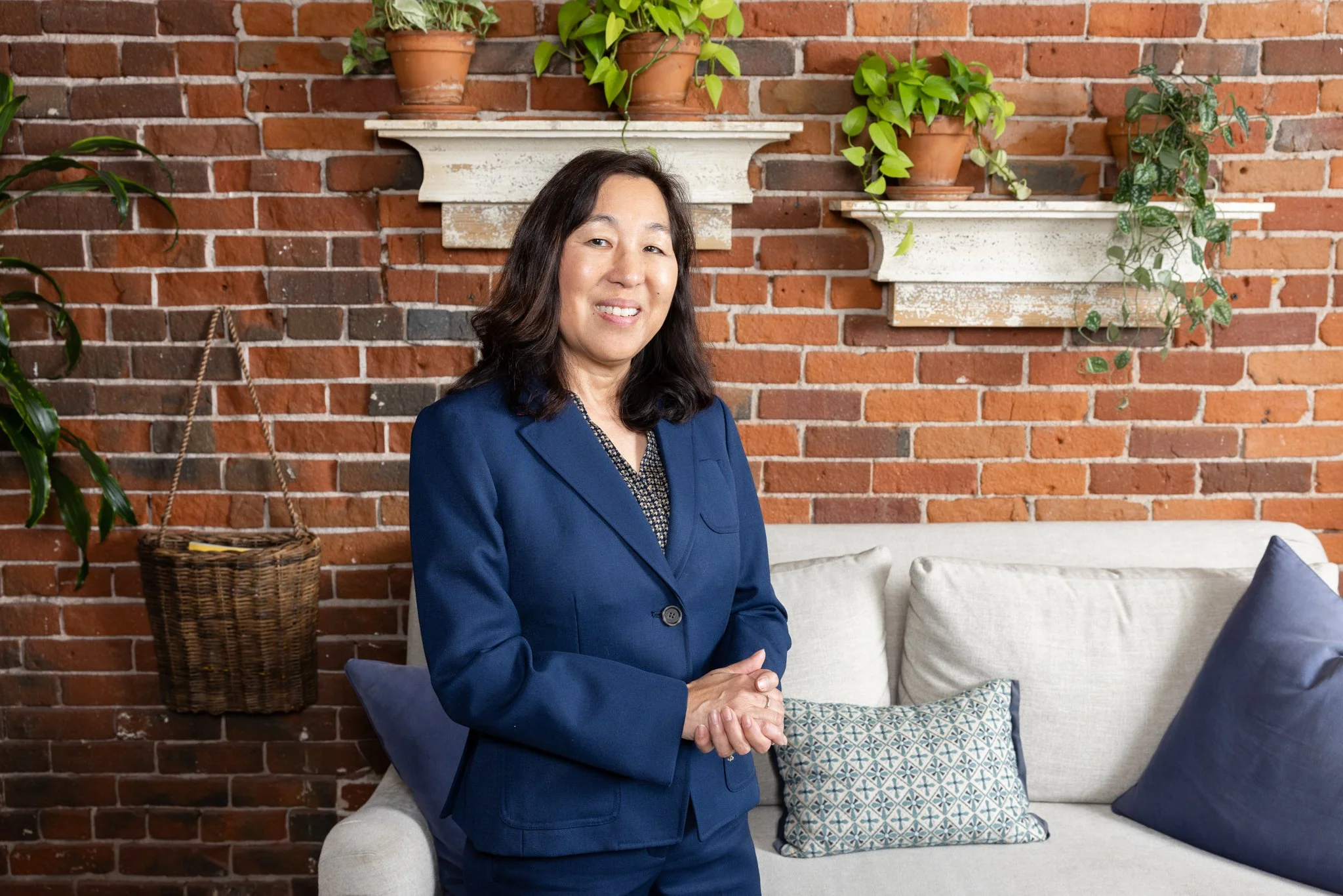 A woman in a blue blazer sitting on a white couch with pillows, indoors, against a brick wall with plants on white shelves.