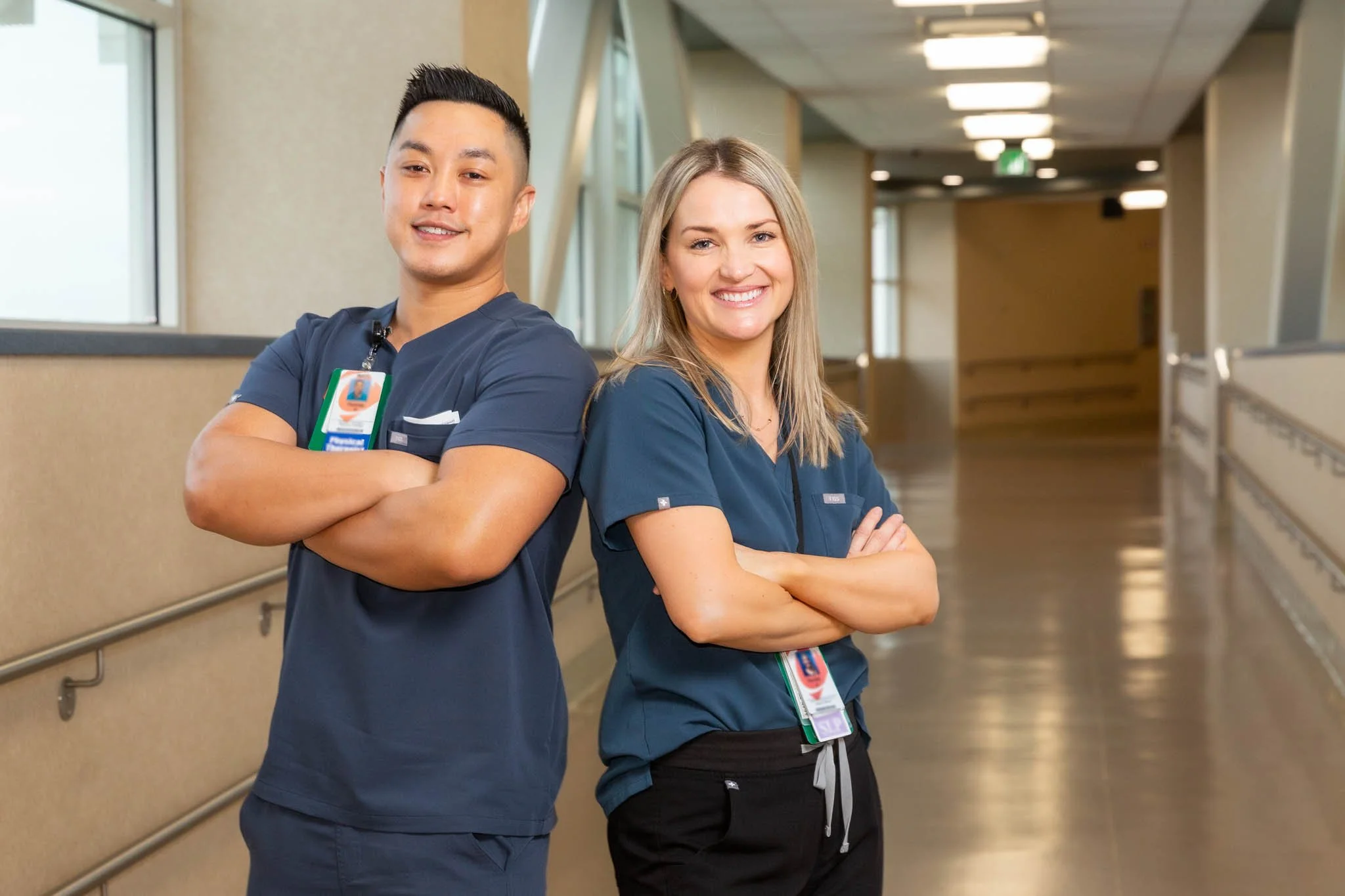 Two healthcare professionals, a man and a woman, standing back to back in a hospital corridor with arms crossed, smiling at the camera. Both are wearing navy blue scrubs and have ID badges.