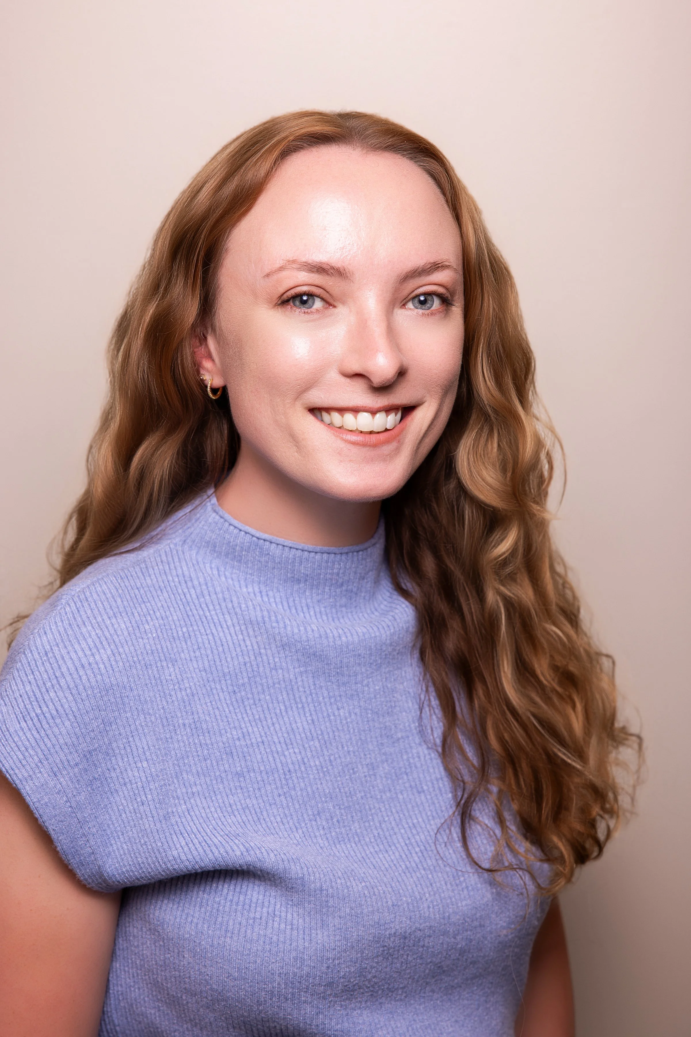 Close-up of a woman with long, curly auburn hair wearing a lavender top, smiling and looking at the camera against a plain beige background.