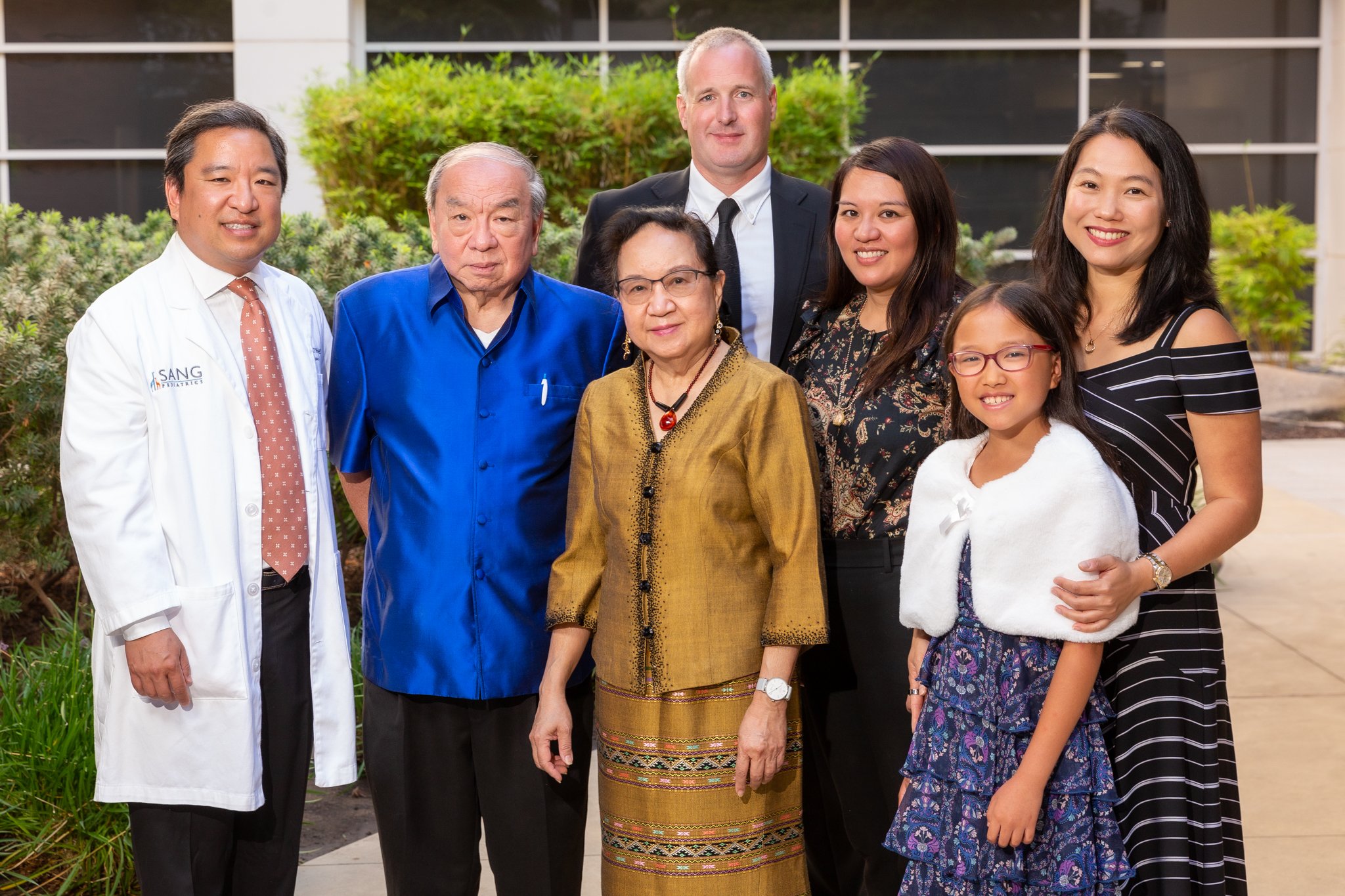 Group of people standing outdoors in front of bushes, including medical professionals, elderly individuals, and a young girl, smiling at the camera.