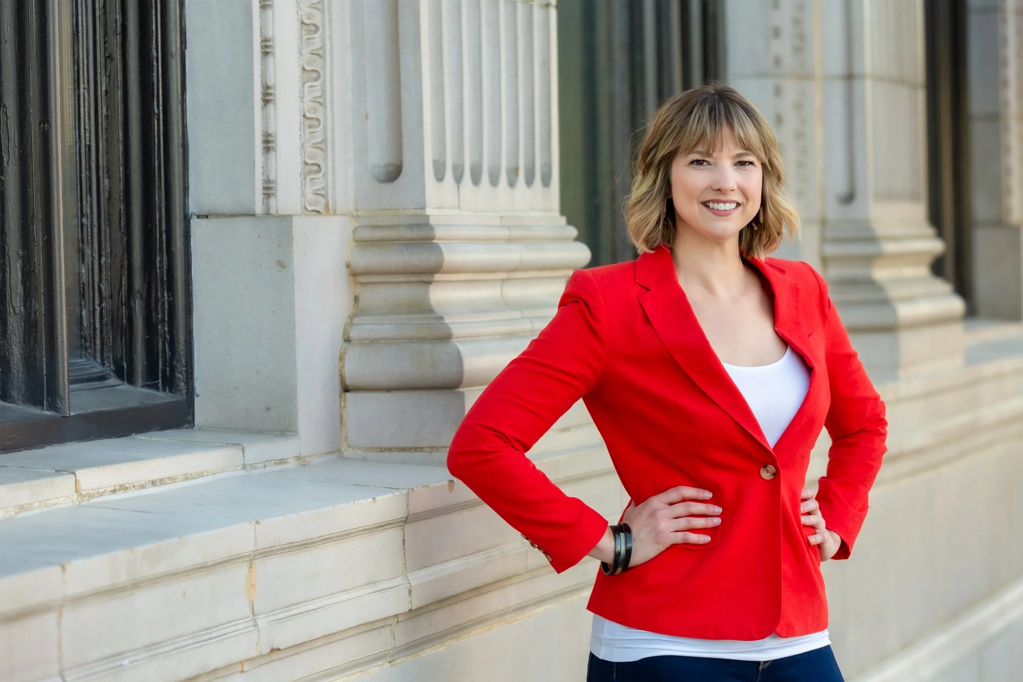 A woman with shoulder-length blond hair wearing a red blazer, white top, and jeans standing outside near columns and black iron fencing, smiling with hands on hips.