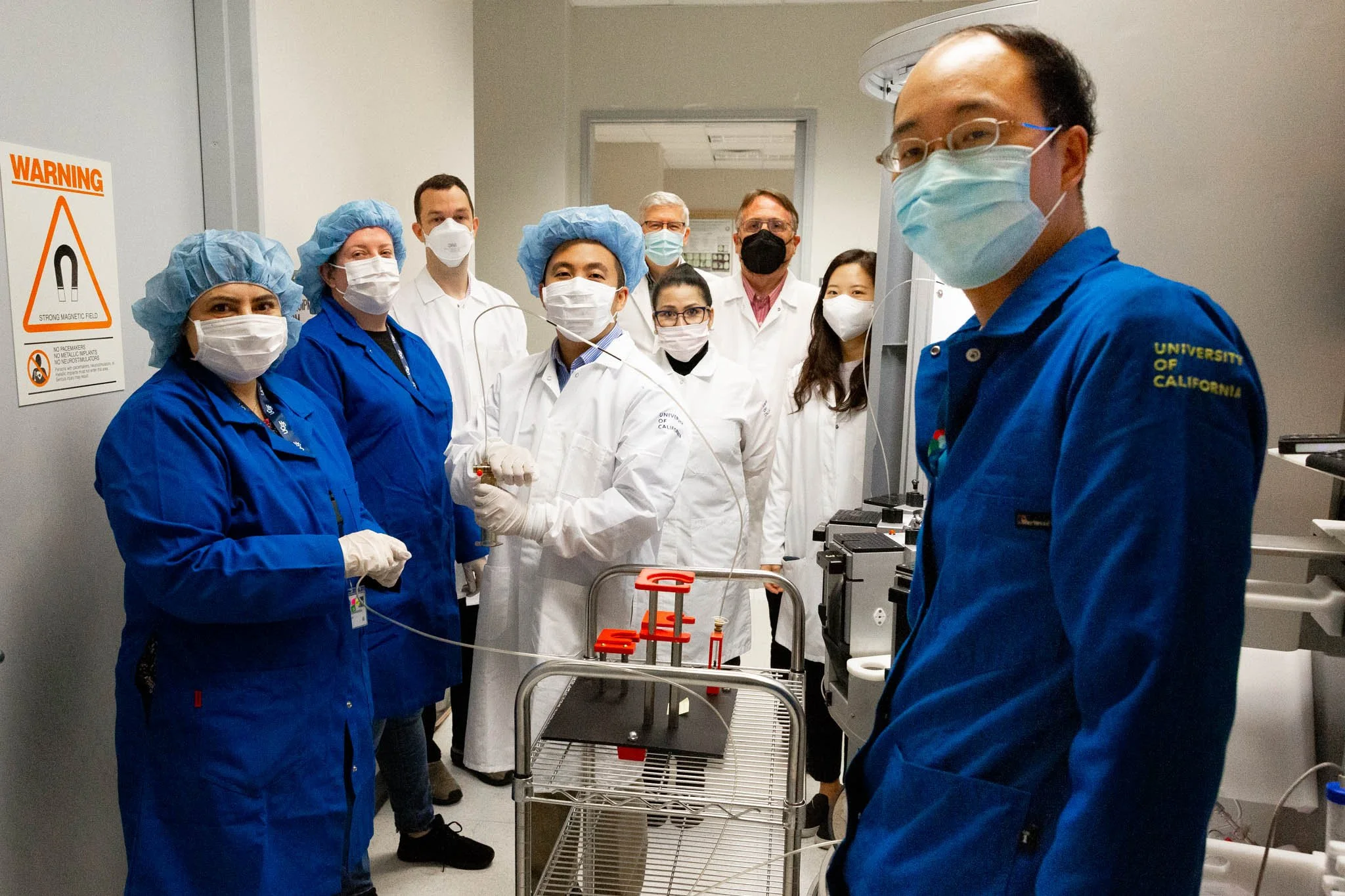 Group of healthcare professionals and researchers in a laboratory wearing masks and blue or white lab coats, standing around equipment.