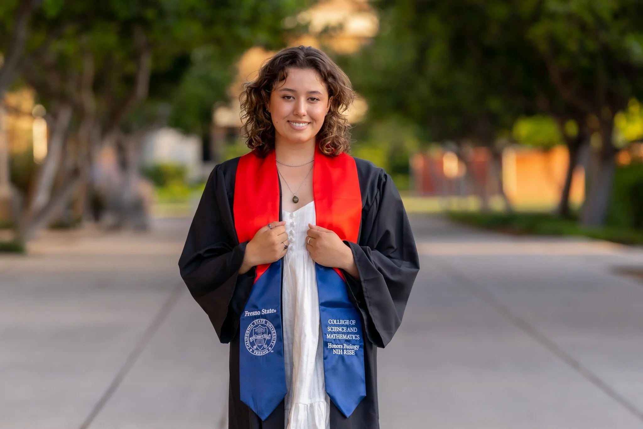 A young woman in a graduation gown and cap, smiling while holding the collar of her gown, standing on a pathway lined with trees.