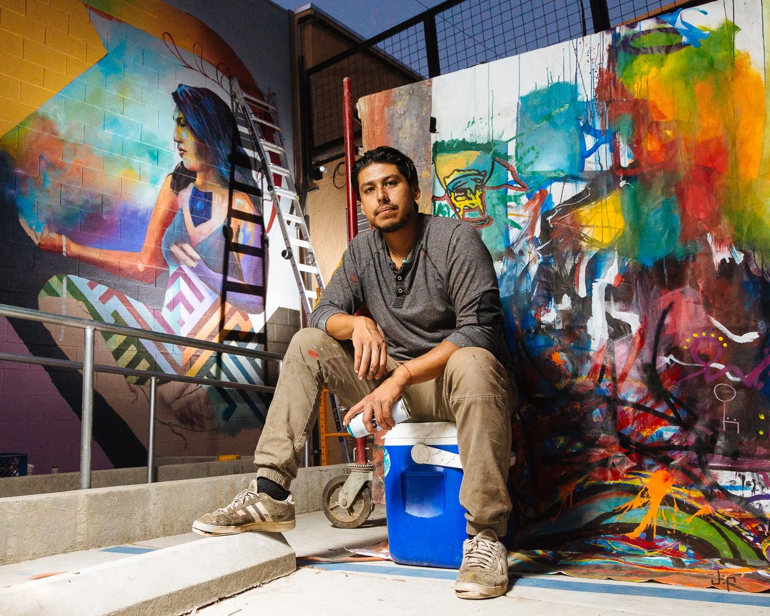 An artist sitting on a cooler in an art studio surrounded by colorful murals and paintings, with ladders and art supplies around.