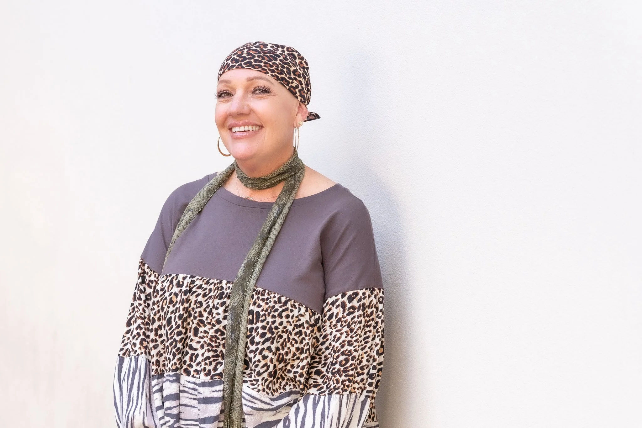 A woman with blonde hair smiling, wearing a leopard print headscarf and a multicolored top with leopard and zebra patterns, standing against a white wall.
