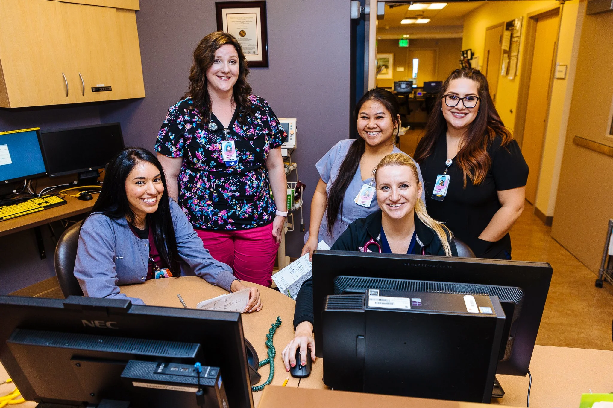 Group of five diverse nurses and healthcare professionals gathered around a desk in a hospital or medical office, smiling at the camera.