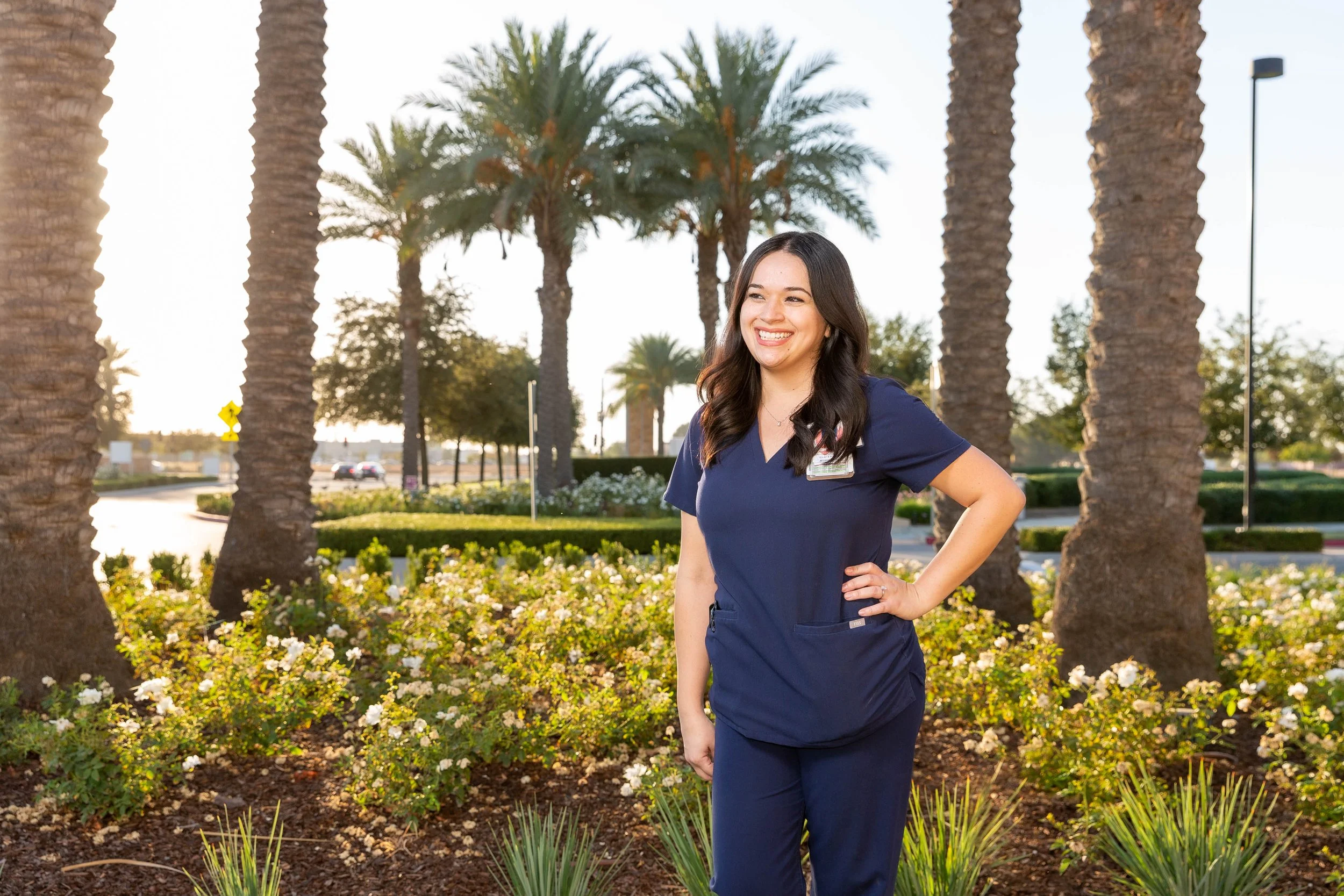 A smiling woman in navy scrubs standing outdoors among palm trees and flowering bushes.