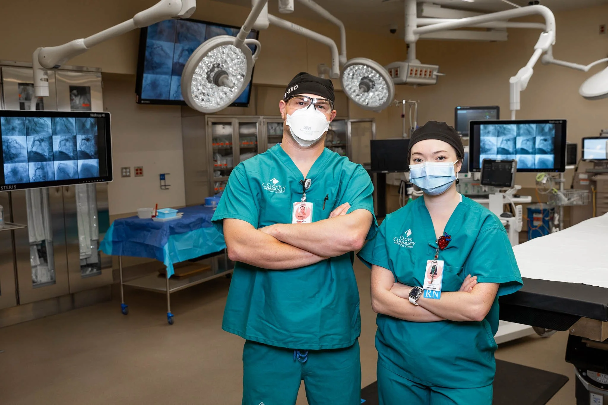 Two medical professionals wearing scrubs and face masks standing in an operating room with medical equipment and monitors in the background.
