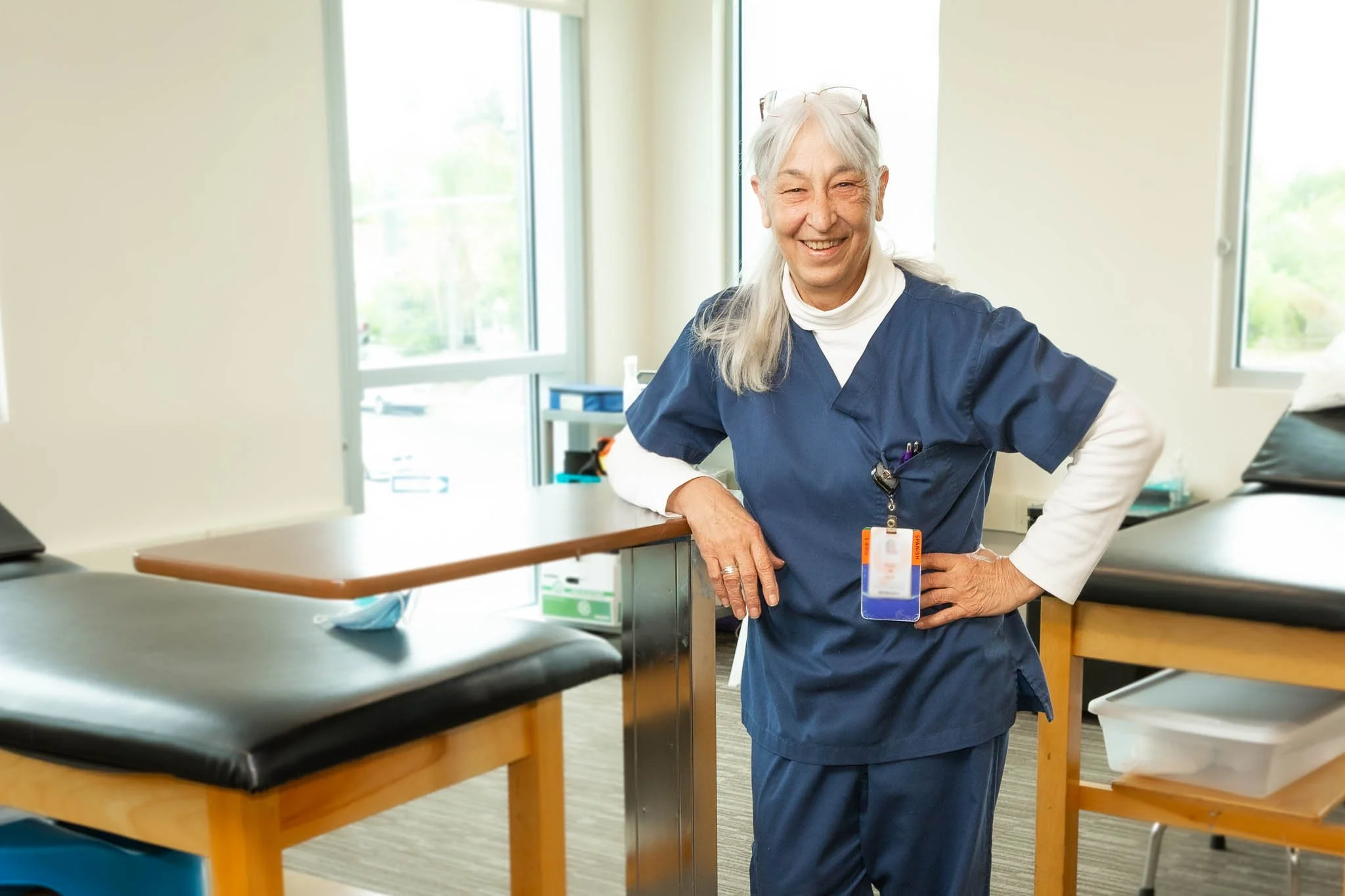 A cheerful healthcare professional with long gray hair, wearing blue scrubs and an ID badge, standing in a medical room with examination tables and large windows.