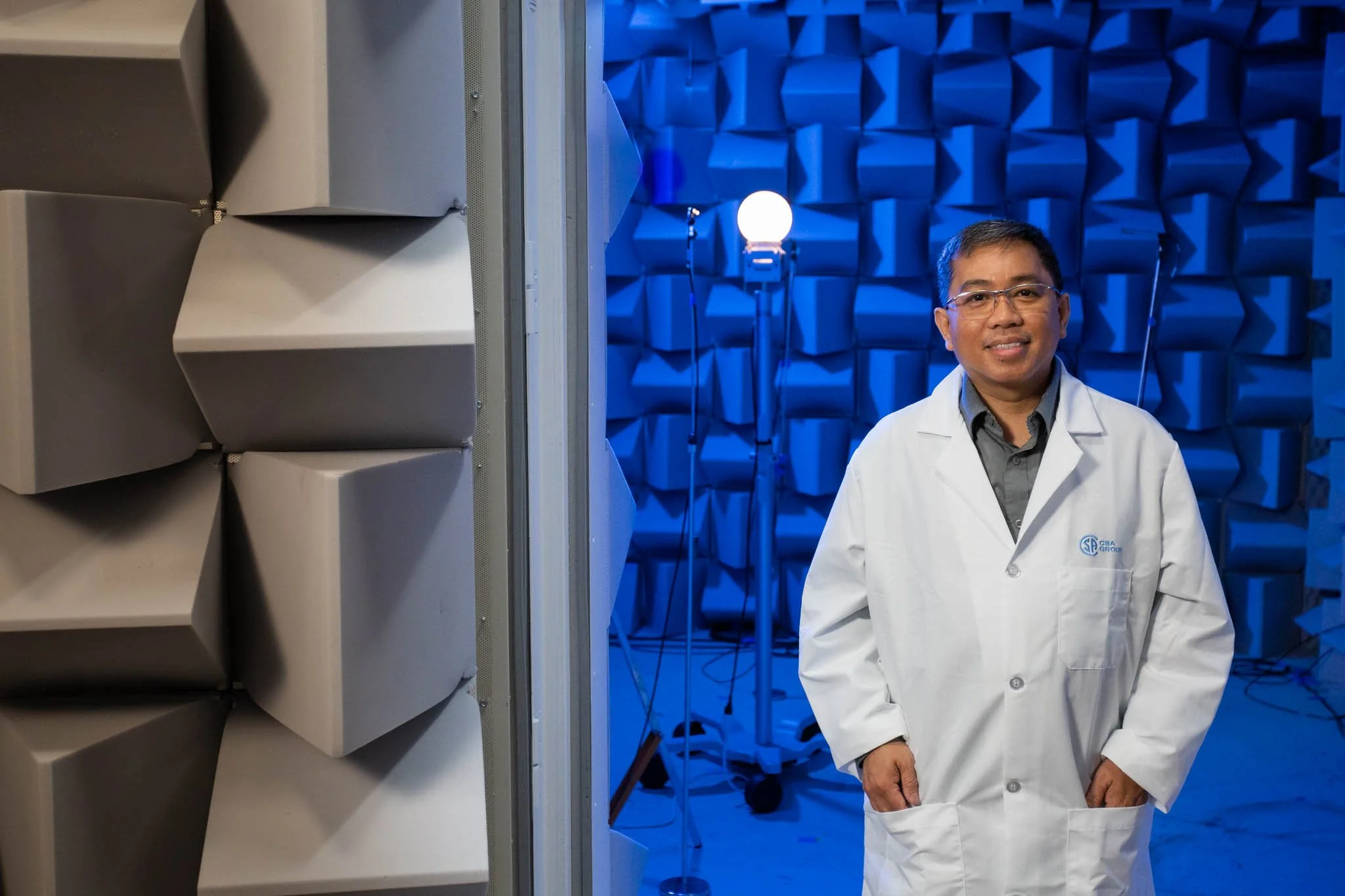 A scientist or engineer in a white lab coat standing in a soundproof testing environment with blue acoustic panels, audio equipment, and lighting.