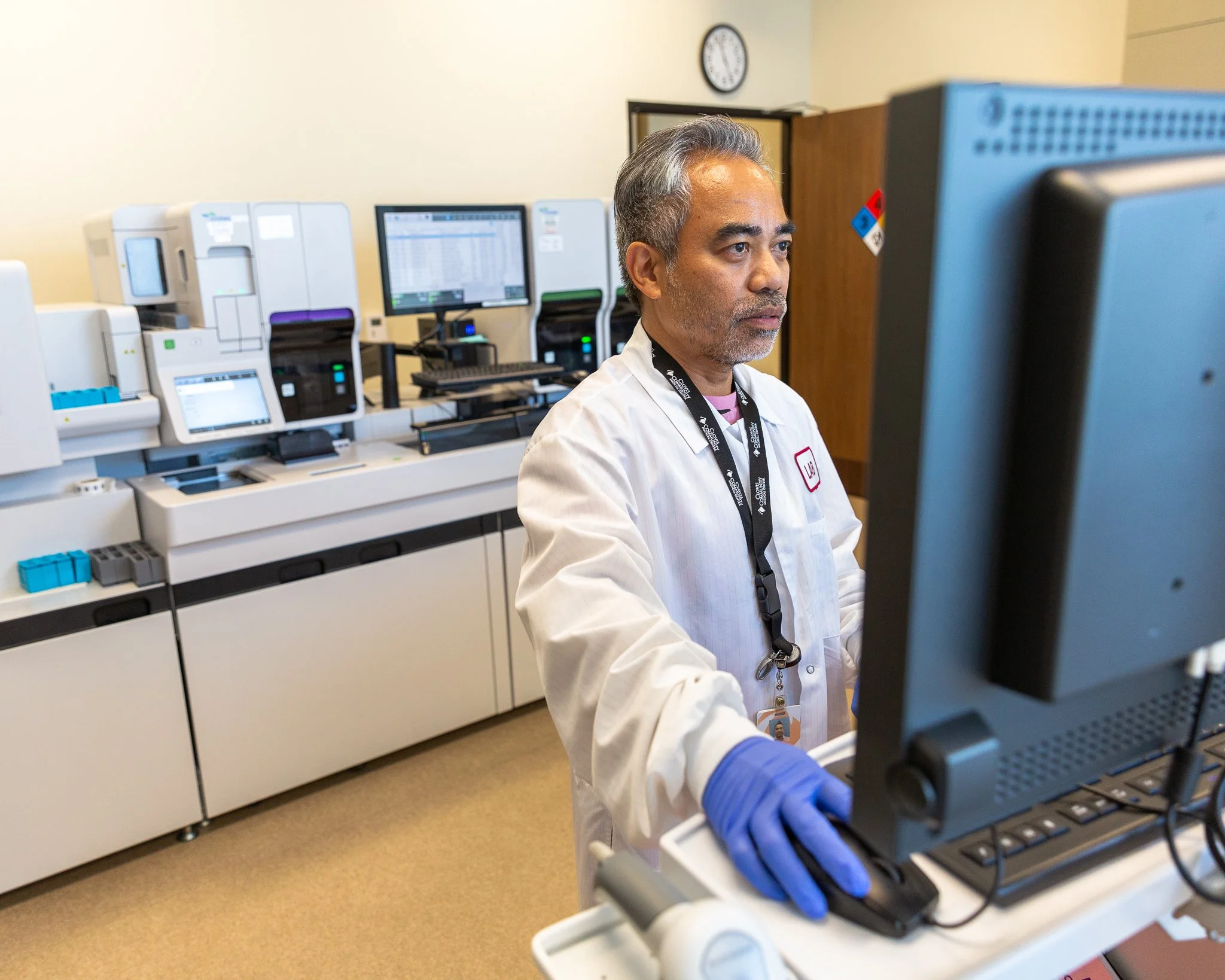 A scientist working at a laboratory computer, wearing a white lab coat and blue gloves, with laboratory equipment and computer monitors in the background.