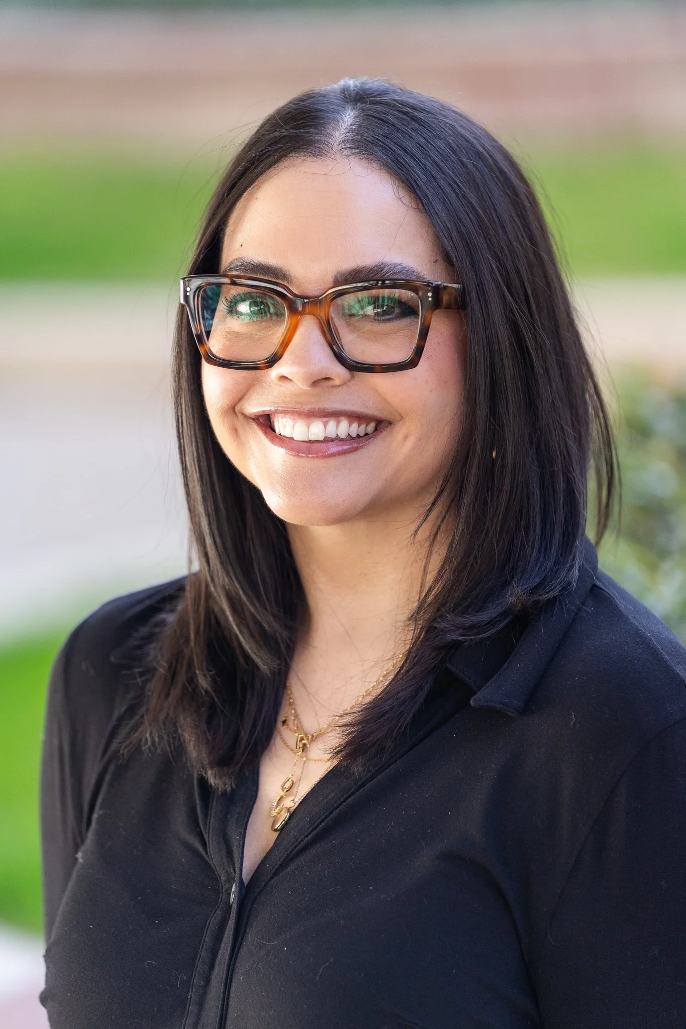 A woman with long dark hair, wearing large tortoiseshell glasses, a black zip-up jacket, and gold jewelry, smiling outdoors with a blurred background of green grass and pathway.