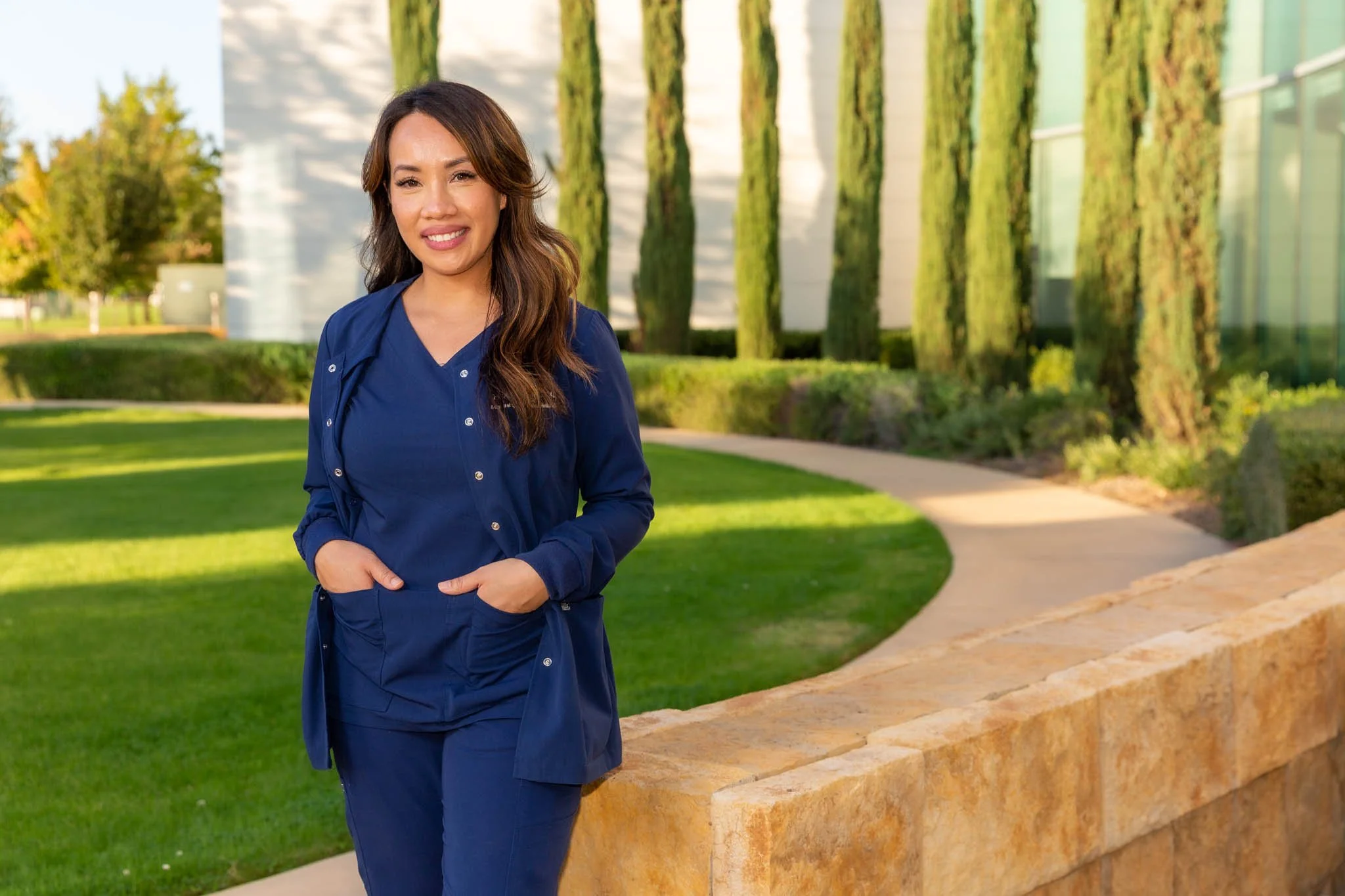 A woman with long brown hair wearing a blue medical uniform, standing outdoors on a sunny day with a green lawn and tall trees in the background.