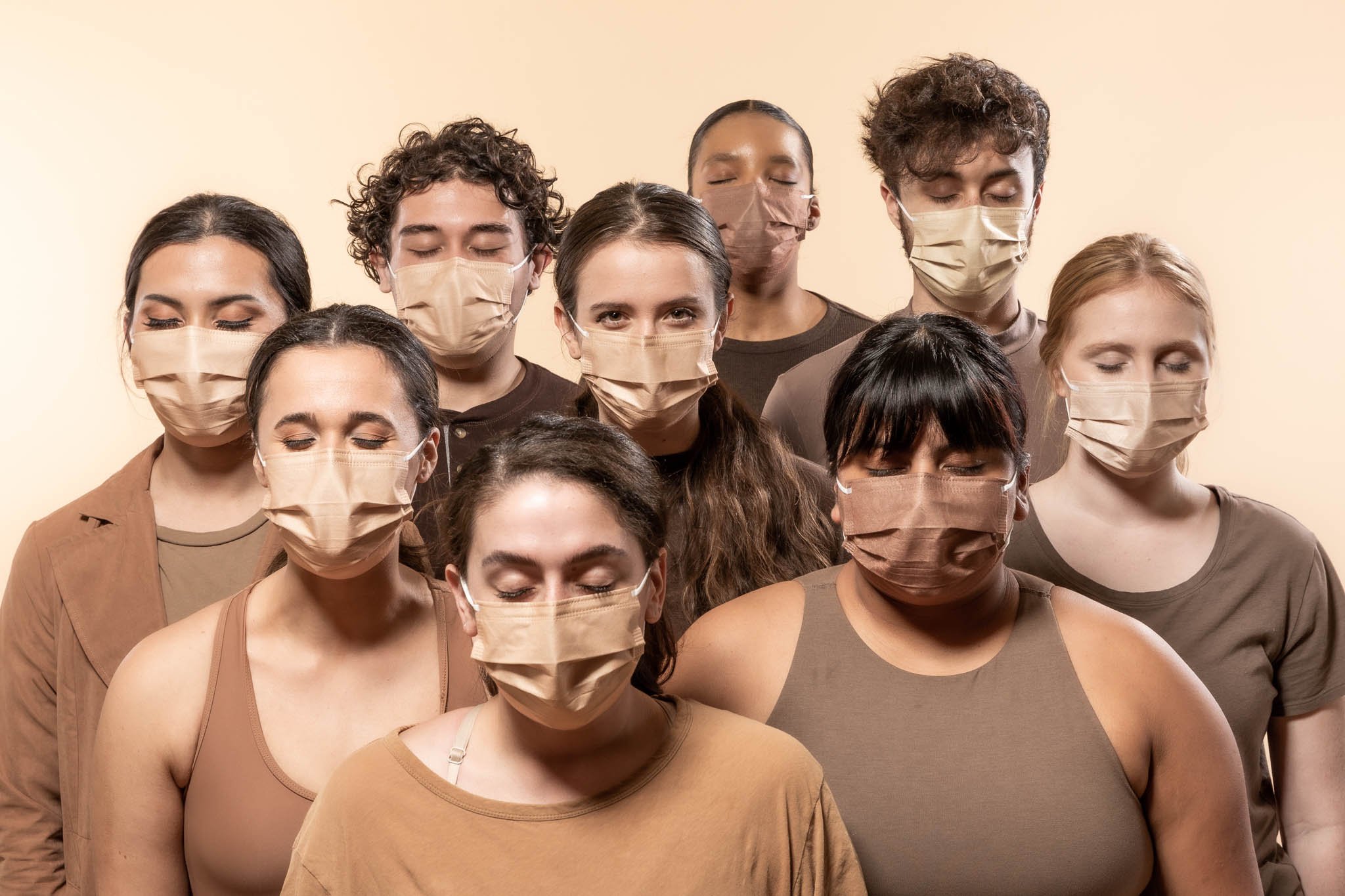 A group of ten women and men standing close together, all wearing beige or brown face masks and with their eyes closed, against a plain background.